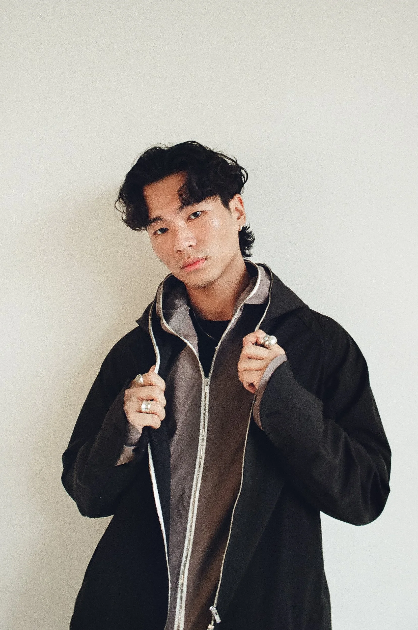 Young man with dark, curly hair wearing a black jacket, holding the collar with both hands, standing against a plain white wall.