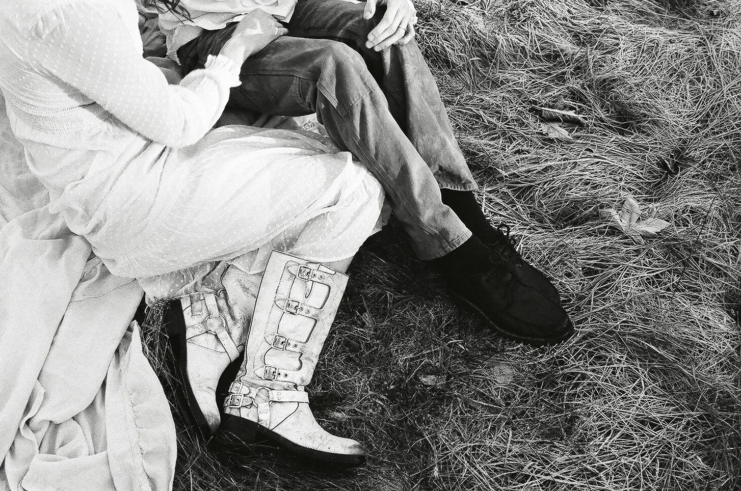 Close-up of two people sitting outdoors on grass, with focus on their legs and shoes, one person wearing white boots with buckles and the other wearing black sneakers.