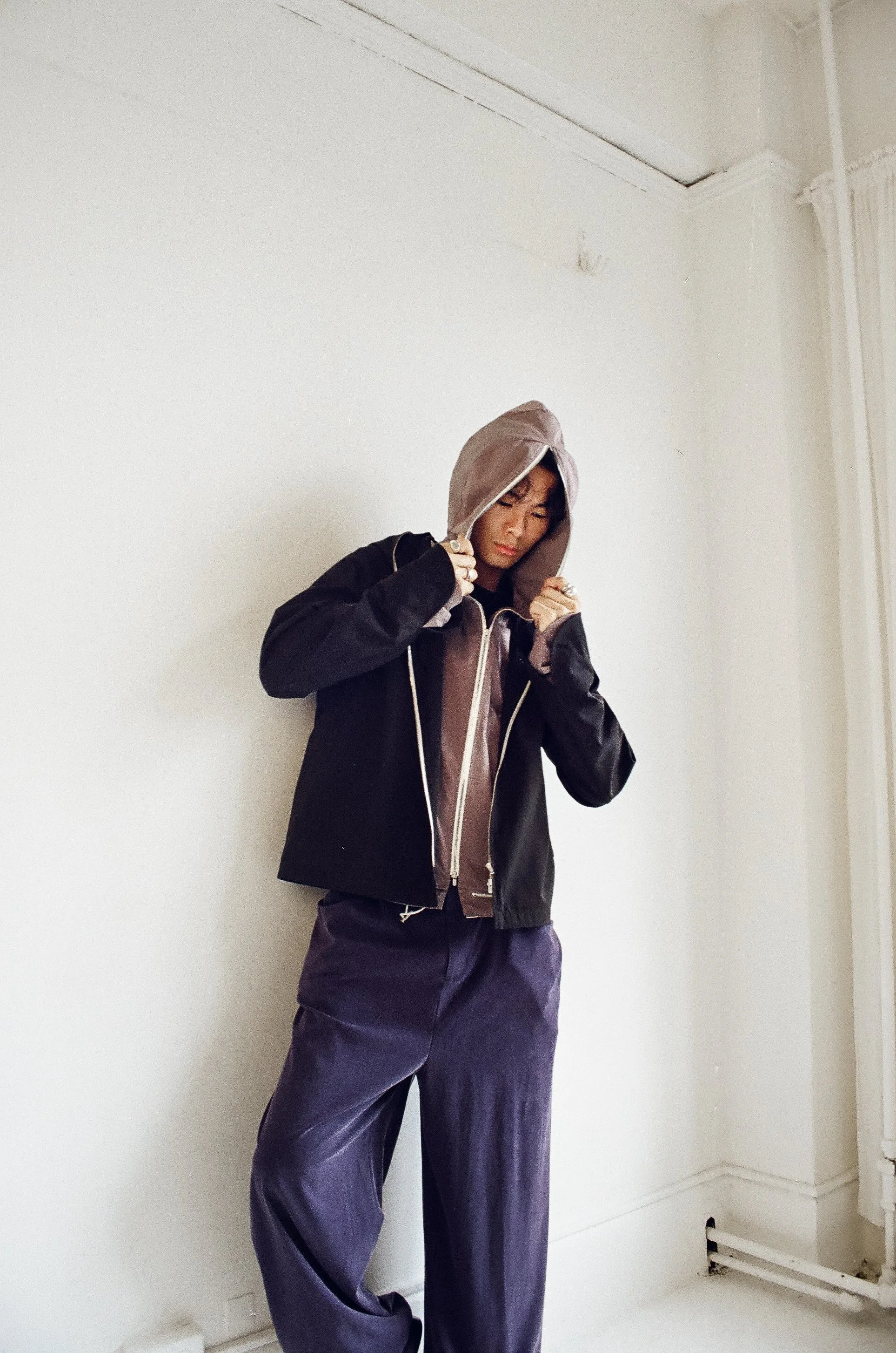 Person wearing a hooded jacket and loose pants, standing against a plain wall, adjusting the jacket's hood.