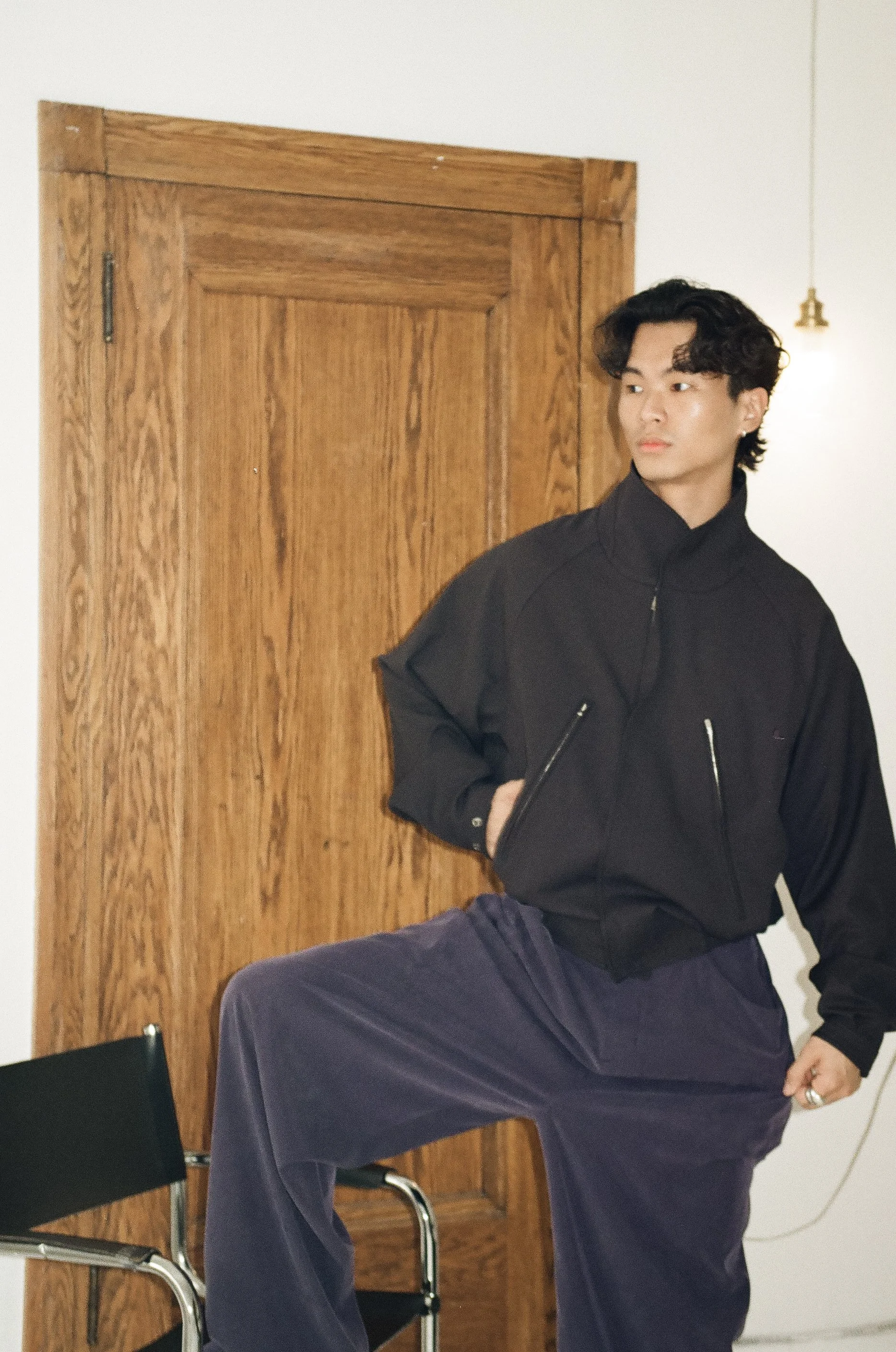 A young man in a black jacket and dark pants poses indoors with one foot elevated on a chair, standing beside a wooden door.