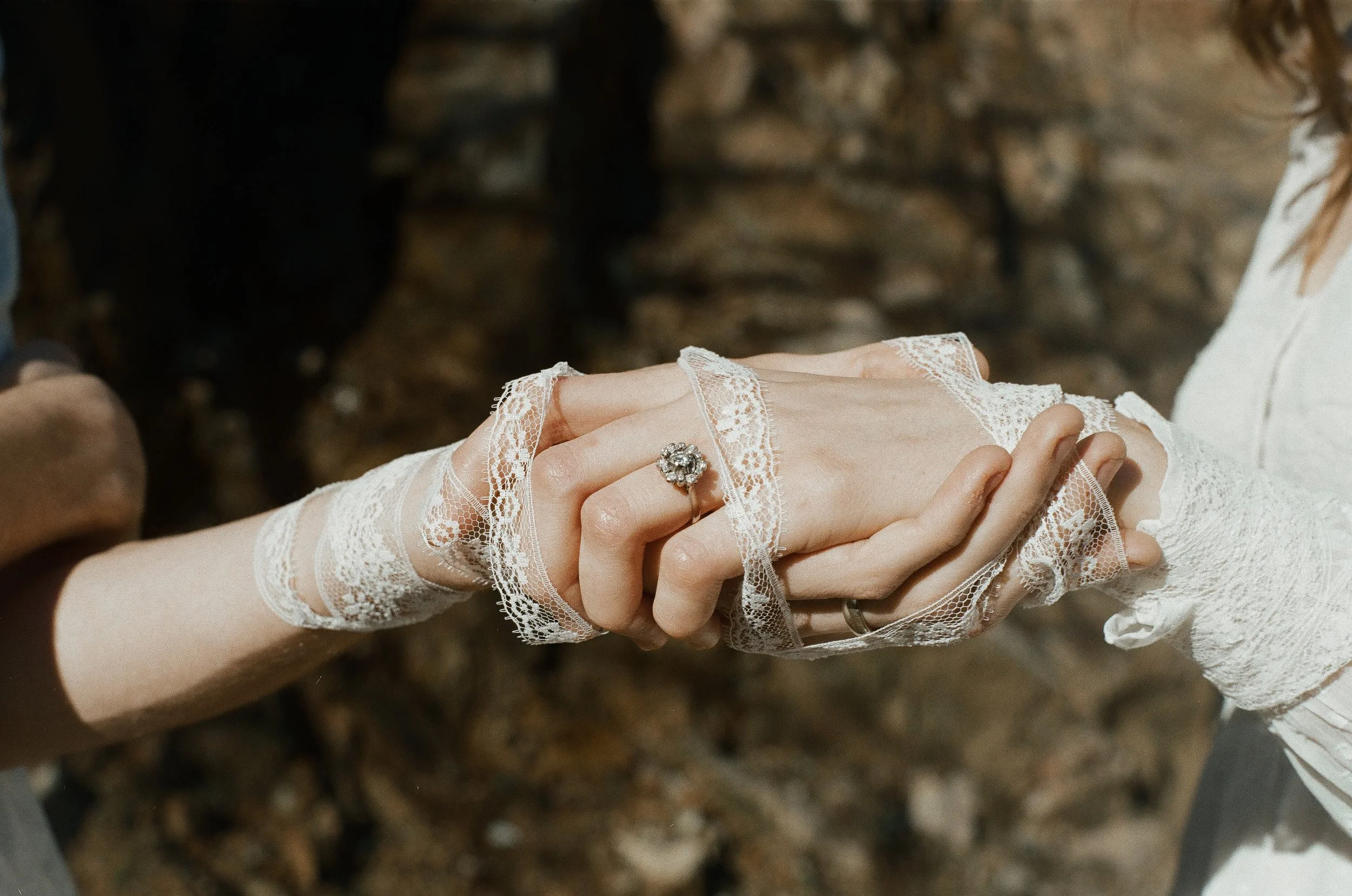 Two people holding hands, one wearing a lace glove and ring, with a background of rocks or soil.