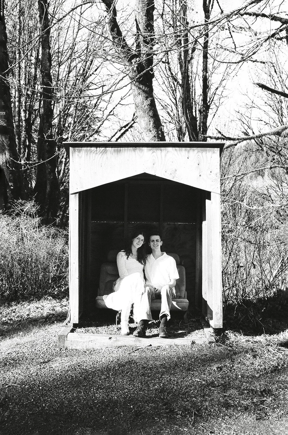 A black and white photo of a couple sitting inside a small wooden shelter outdoors, surrounded by trees with bare branches, suggesting winter.