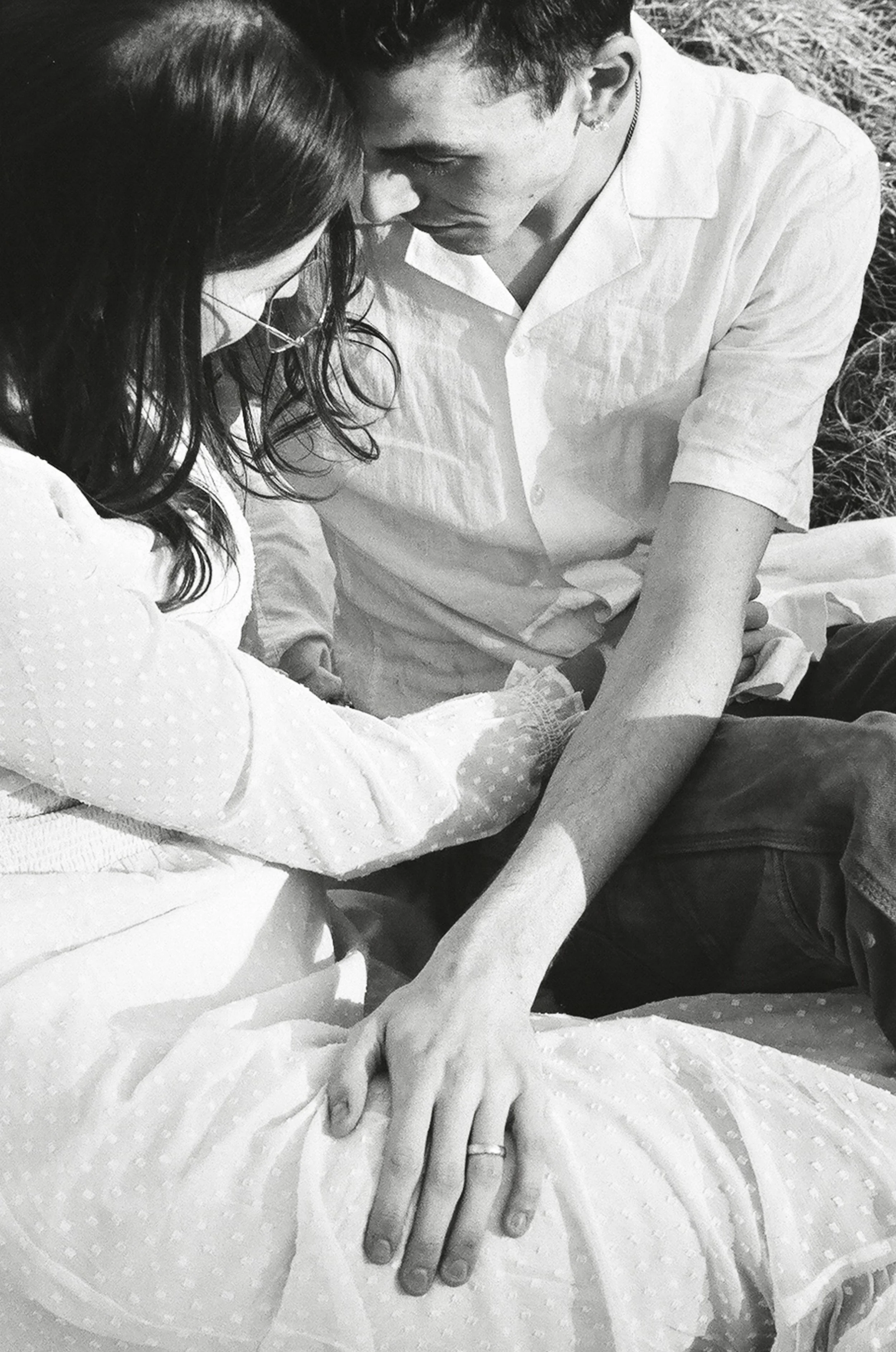 A black and white photo of a couple in an outdoor setting, holding hands and leaning their foreheads together, showing emotional intimacy.