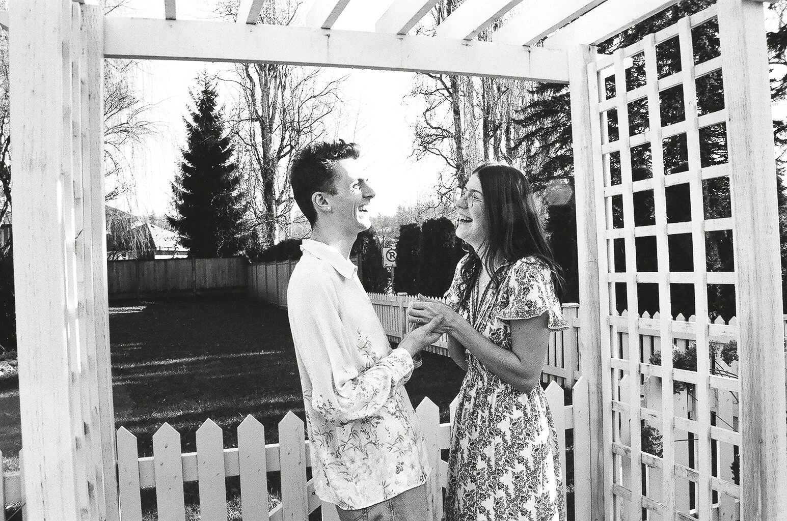 A black-and-white photo of a couple standing outdoors in a white wooden garden archway, smiling at each other, with trees and a fence in the background.