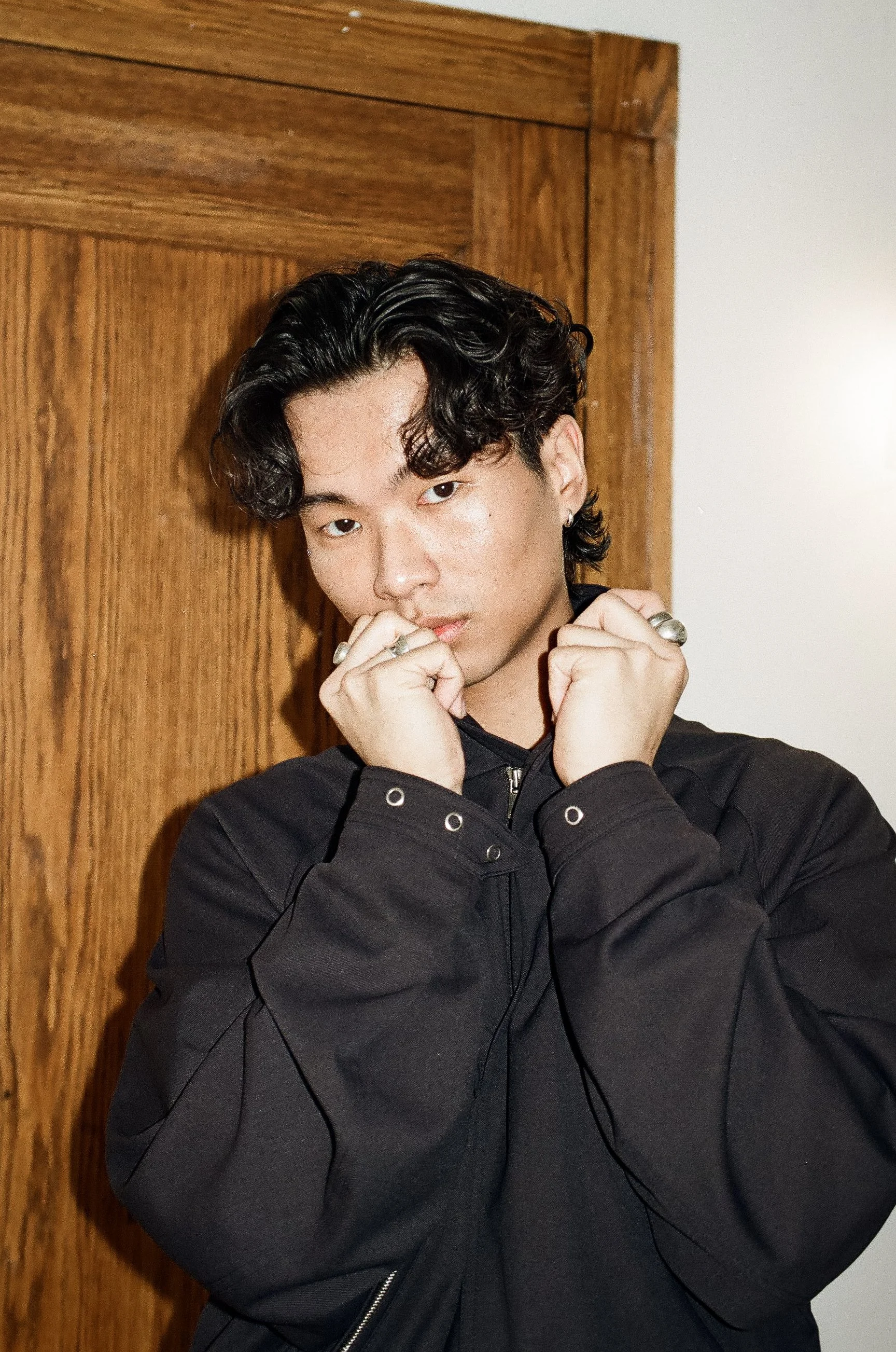 A young man with dark, wavy hair posing indoors in front of a wooden door. He is wearing a black jacket and has rings on both hands, which he is holding close to his face.