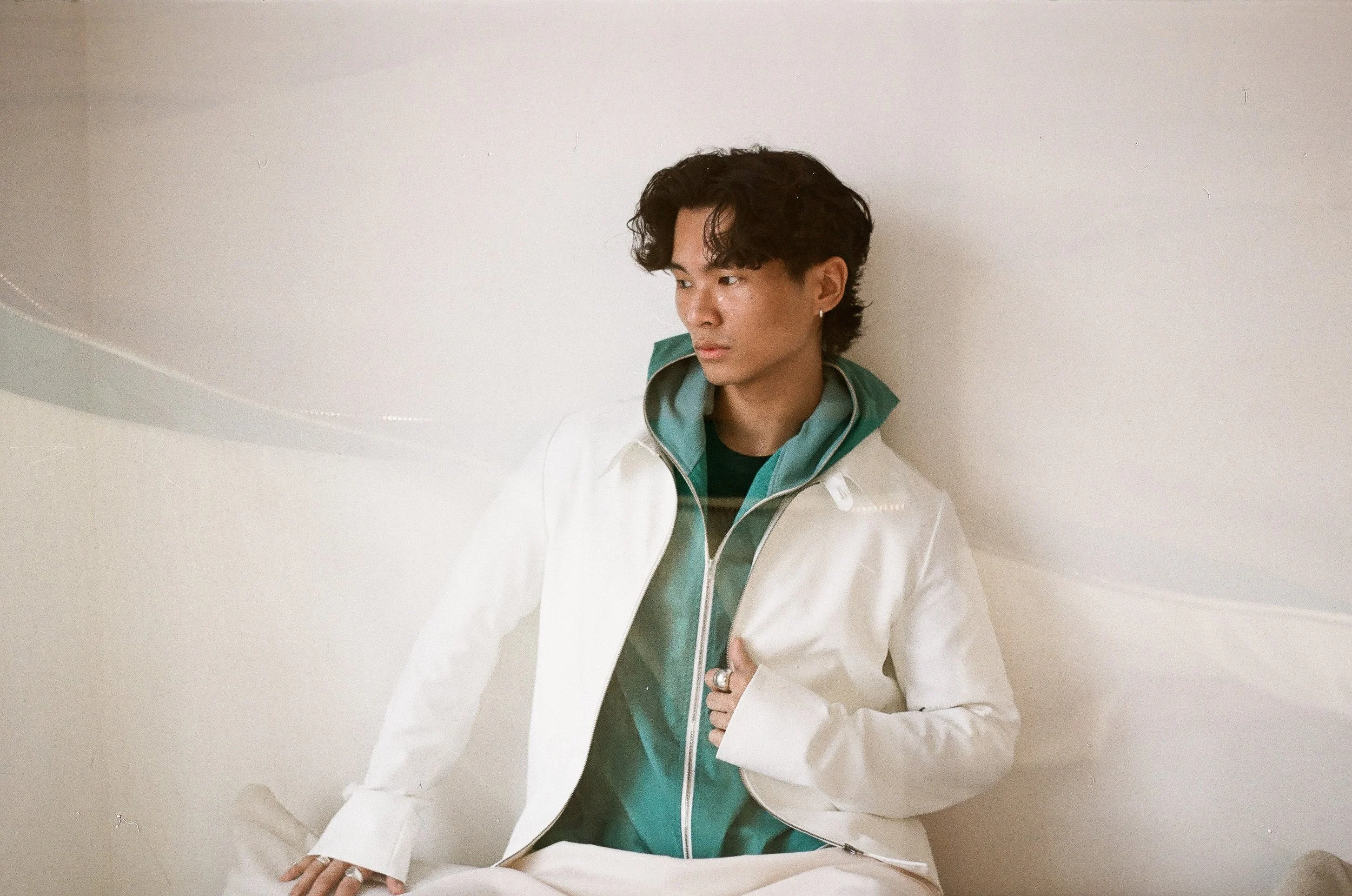 A young man with curly dark hair wearing a white jacket over a green hoodie, sitting against a plain white wall, looking to the side.