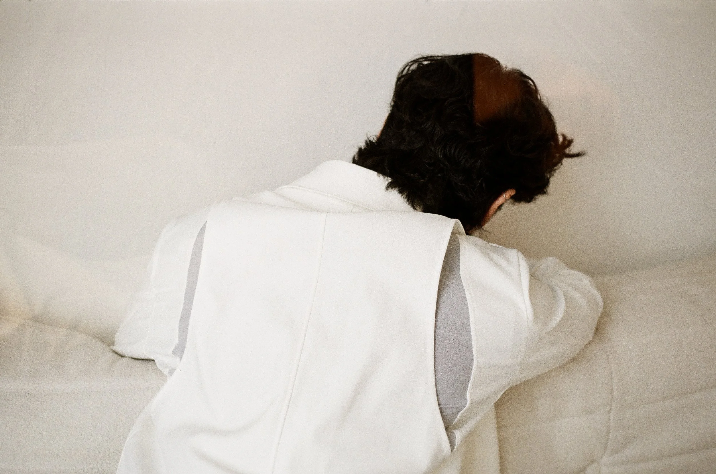 A person with dark, wavy hair sitting on a beige sofa, facing away from the camera, in a white shirt with rolled-up sleeves.