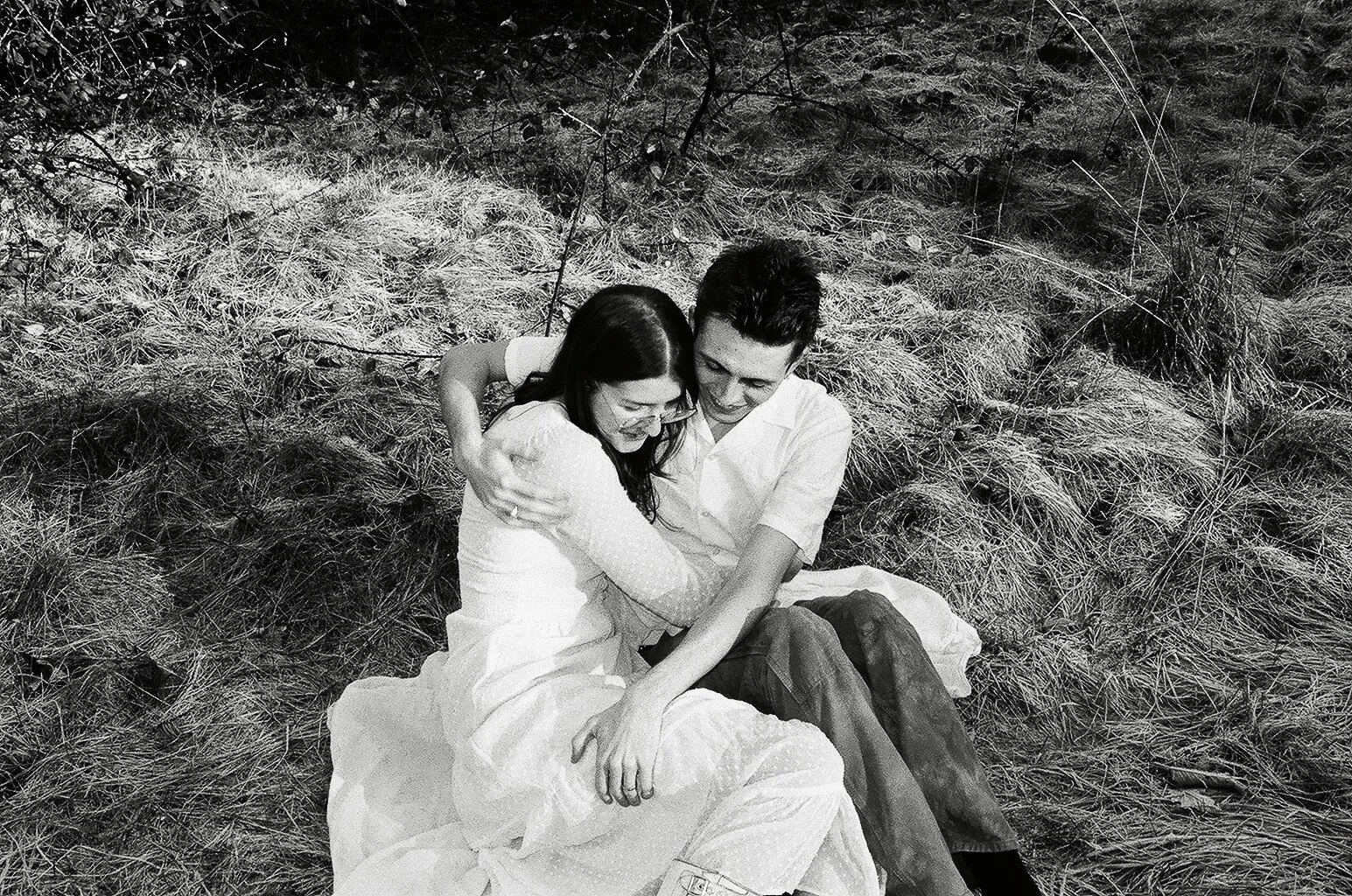 A black and white photo of a couple sitting on the grass, embracing and sharing a joyful moment.