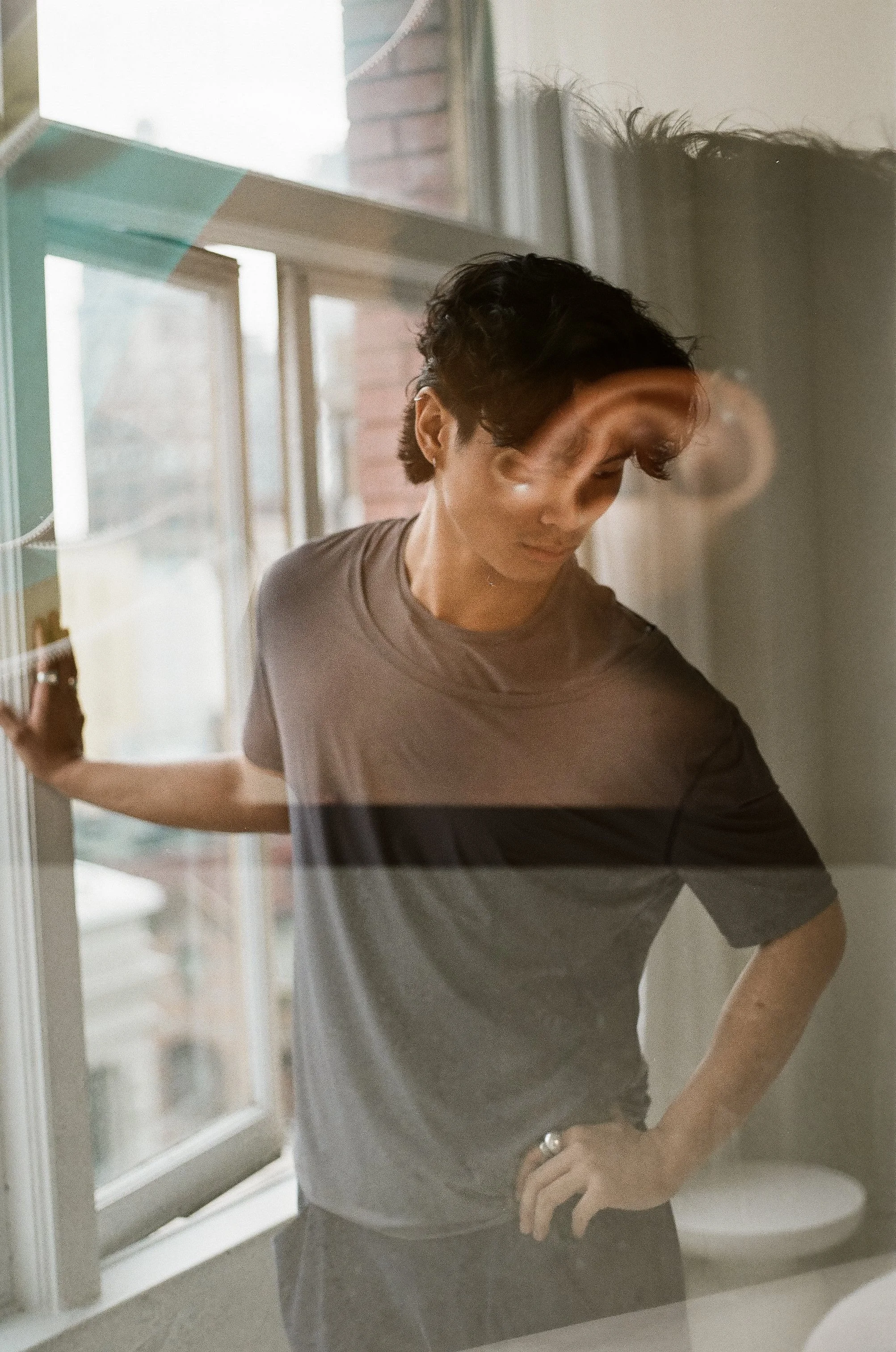 A person with short dark hair is seen through a glass window, wearing a gray T-shirt, leaning against the window frame with one arm, and the other hand on their hip, looking down.