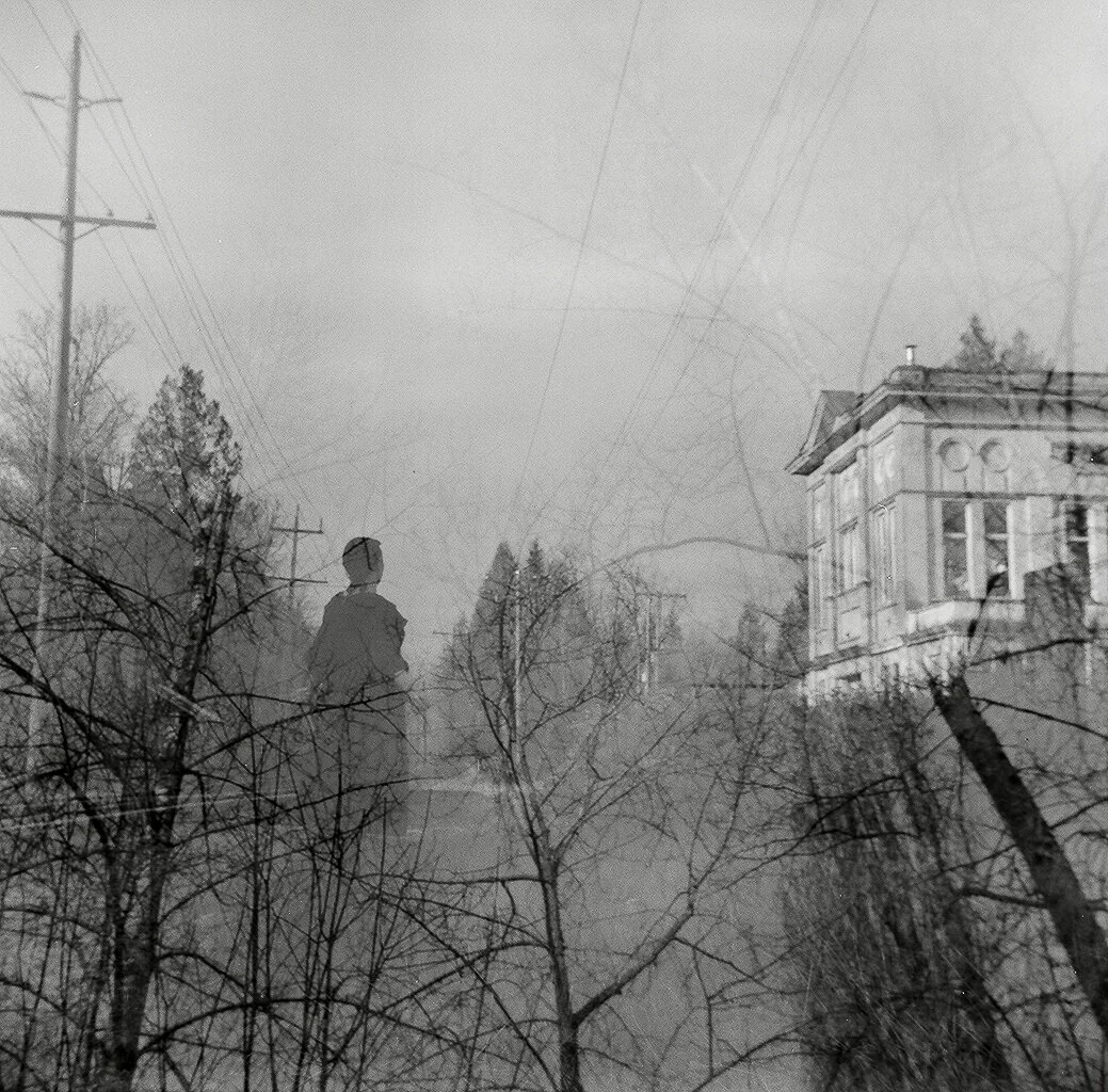 A double exposure photograph blending a house, leafless trees, power lines, and a person standing outdoors.