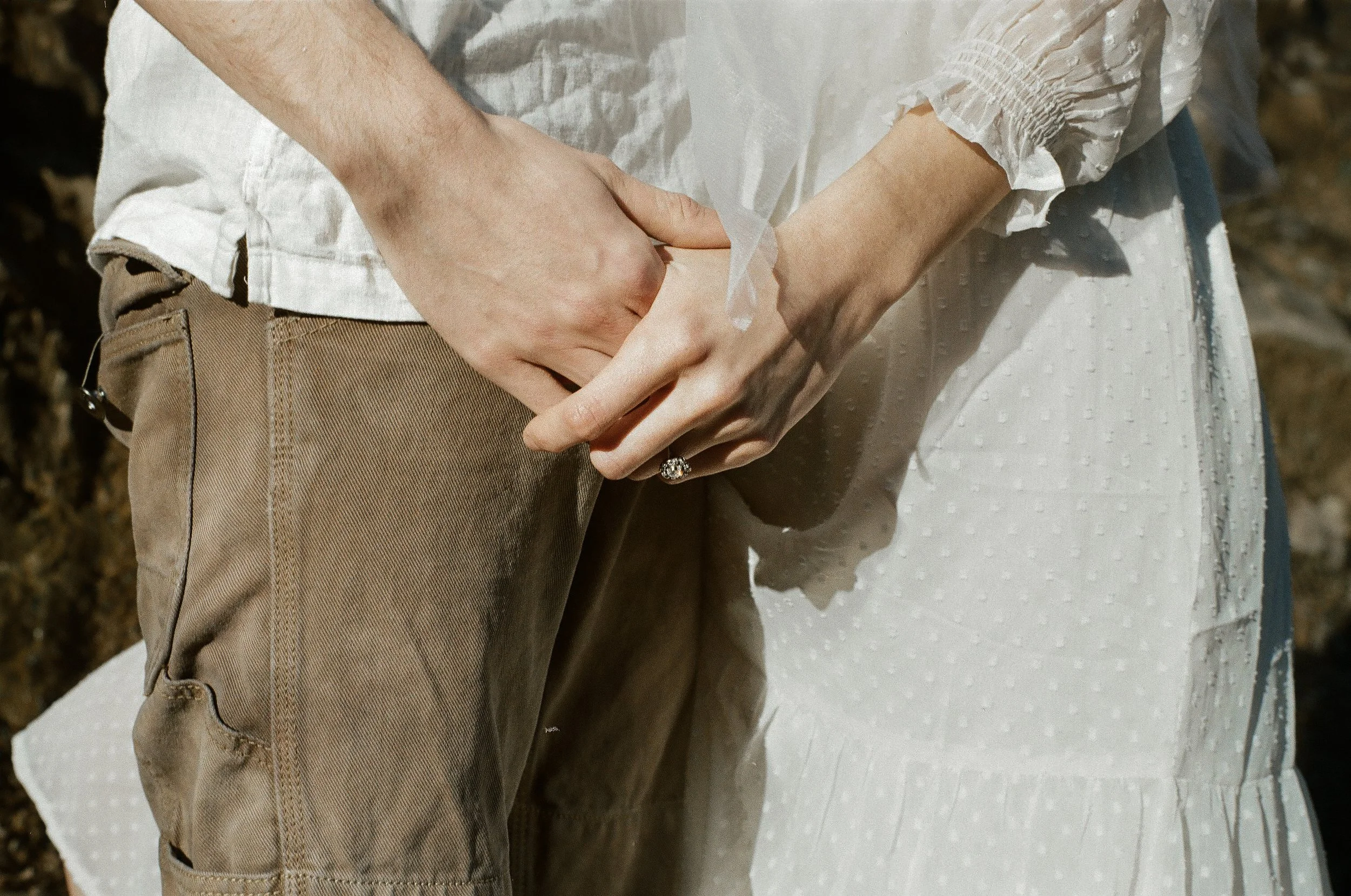 A person wearing a white blouse and beige pants is sitting on a white cushioned surface outdoors, with their hands clasped together in their lap. The person's left hand has a ring on the ring finger.