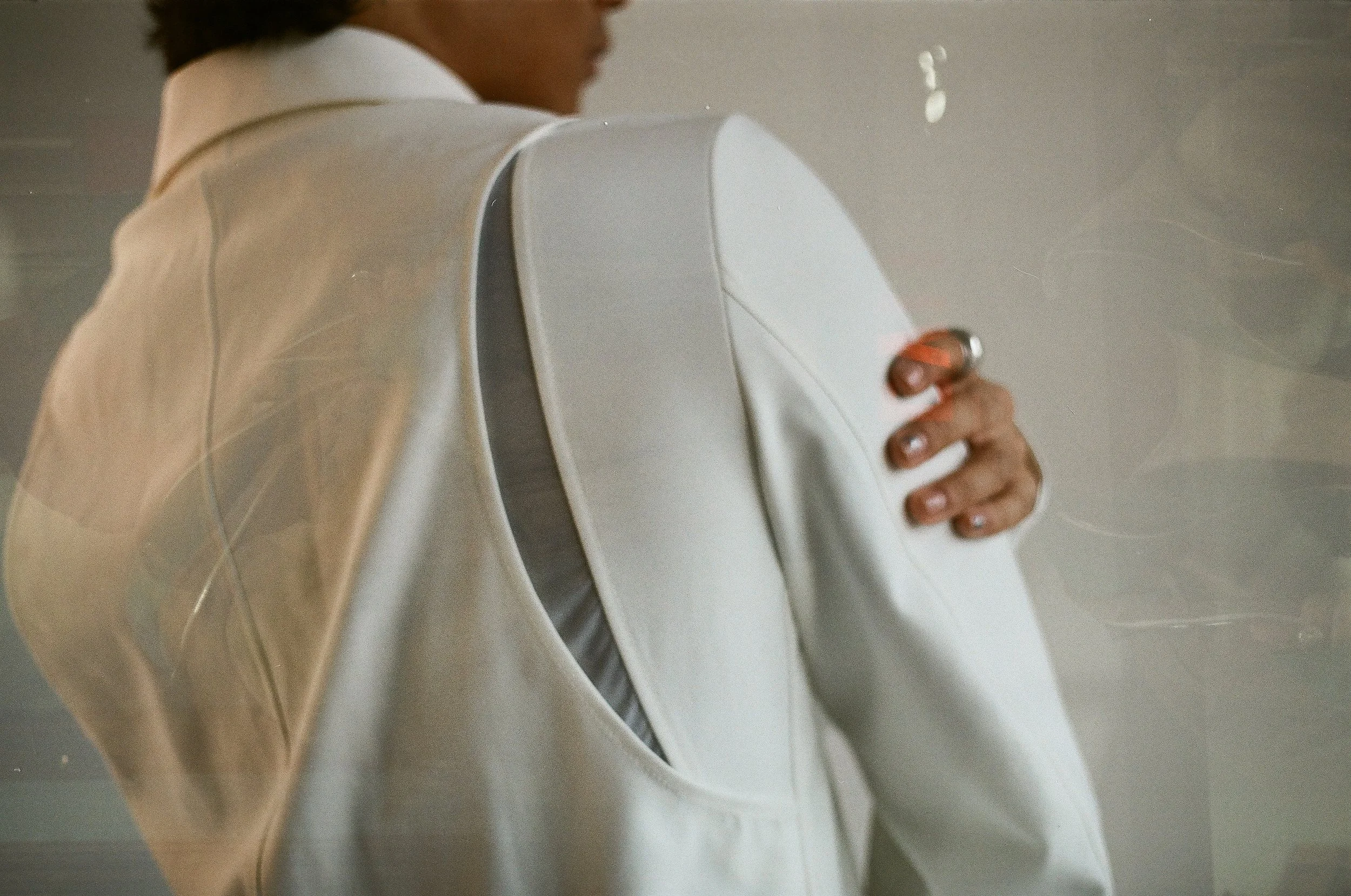 Person wearing a beige jacket and white blazer, holding their arm with their hand.