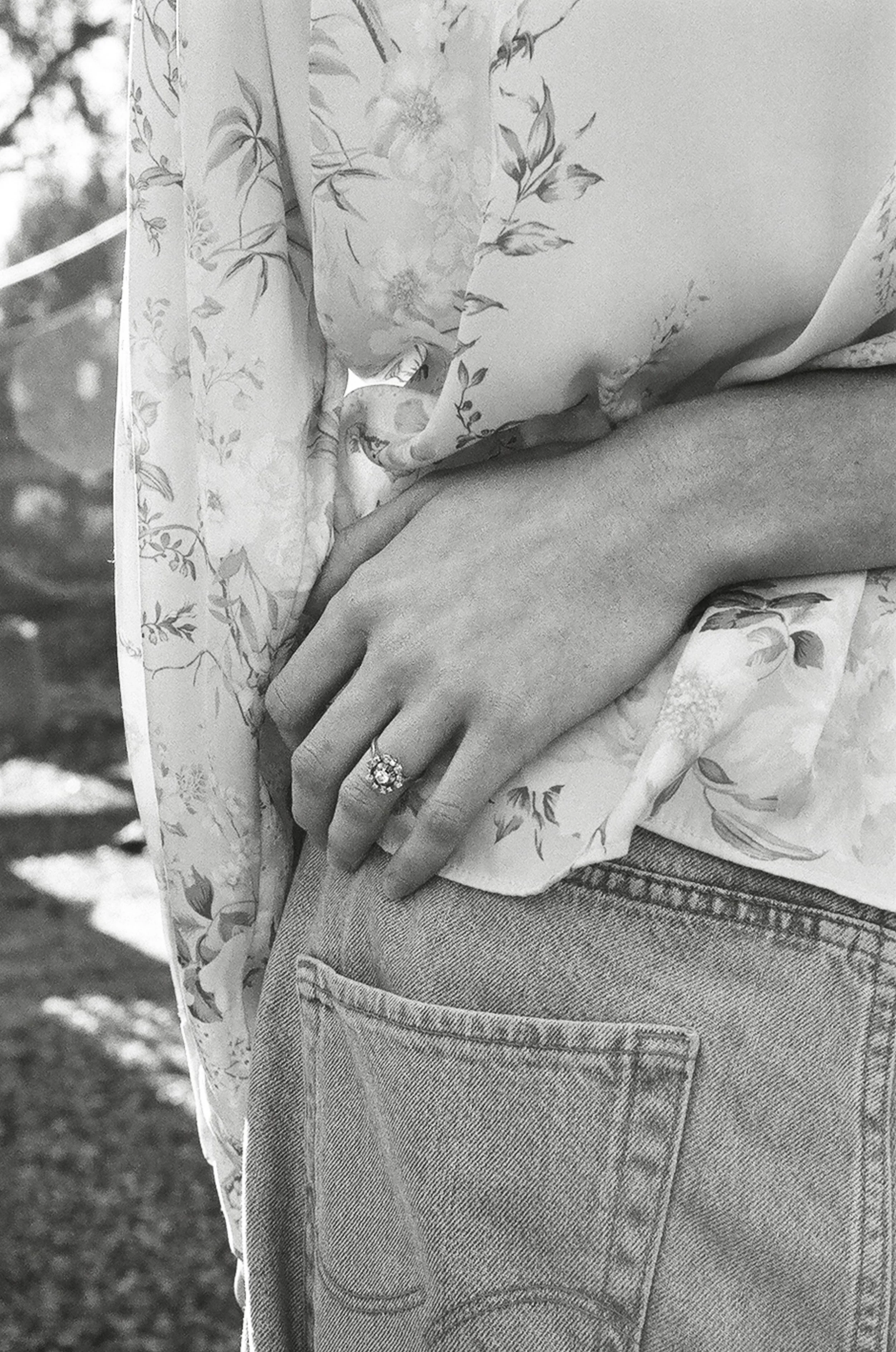 Close-up of a person wearing floral-patterned clothing and jeans, with their left hand resting on their hip, showing an engagement ring on the ring finger.