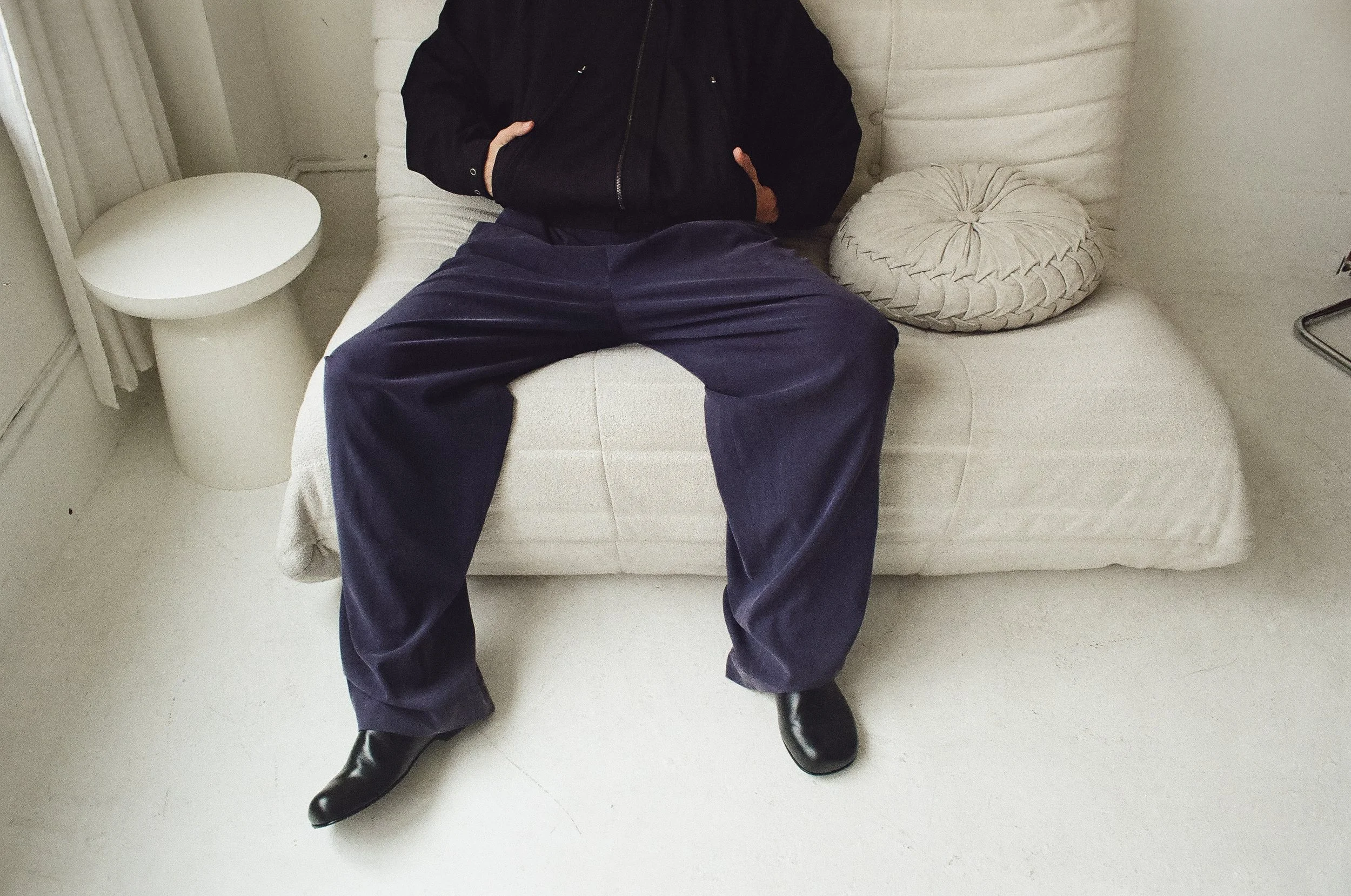 Person sitting on a white sofa with black leather shoes, wearing dark blue pants and a black jacket. There is a round pillow on the sofa and a white side table next to it.