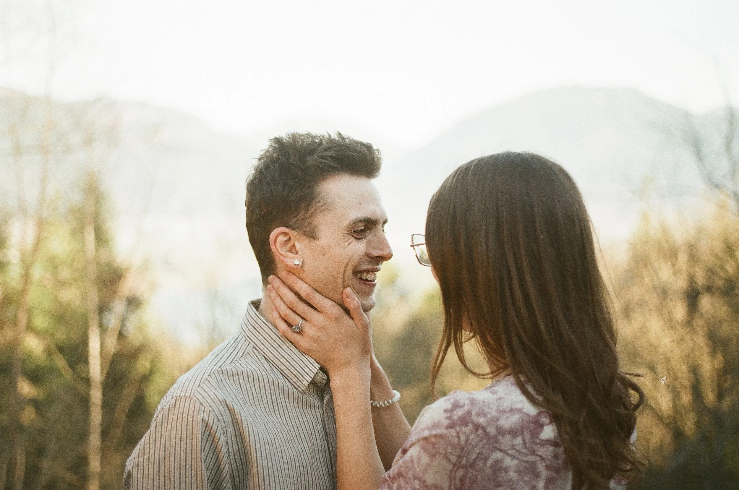 A man and woman face to face outdoors, smiling, with the woman holding the man's face gently.
