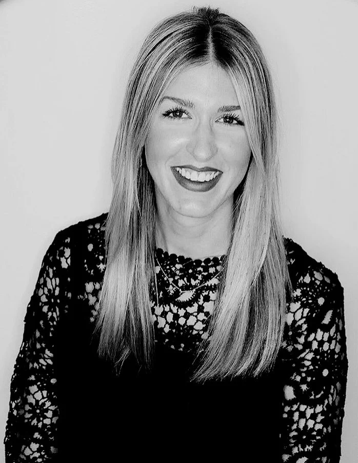 A young woman with straight, shoulder-length hair and makeup, smiling with teeth showing, wearing a black lace top against a plain background.