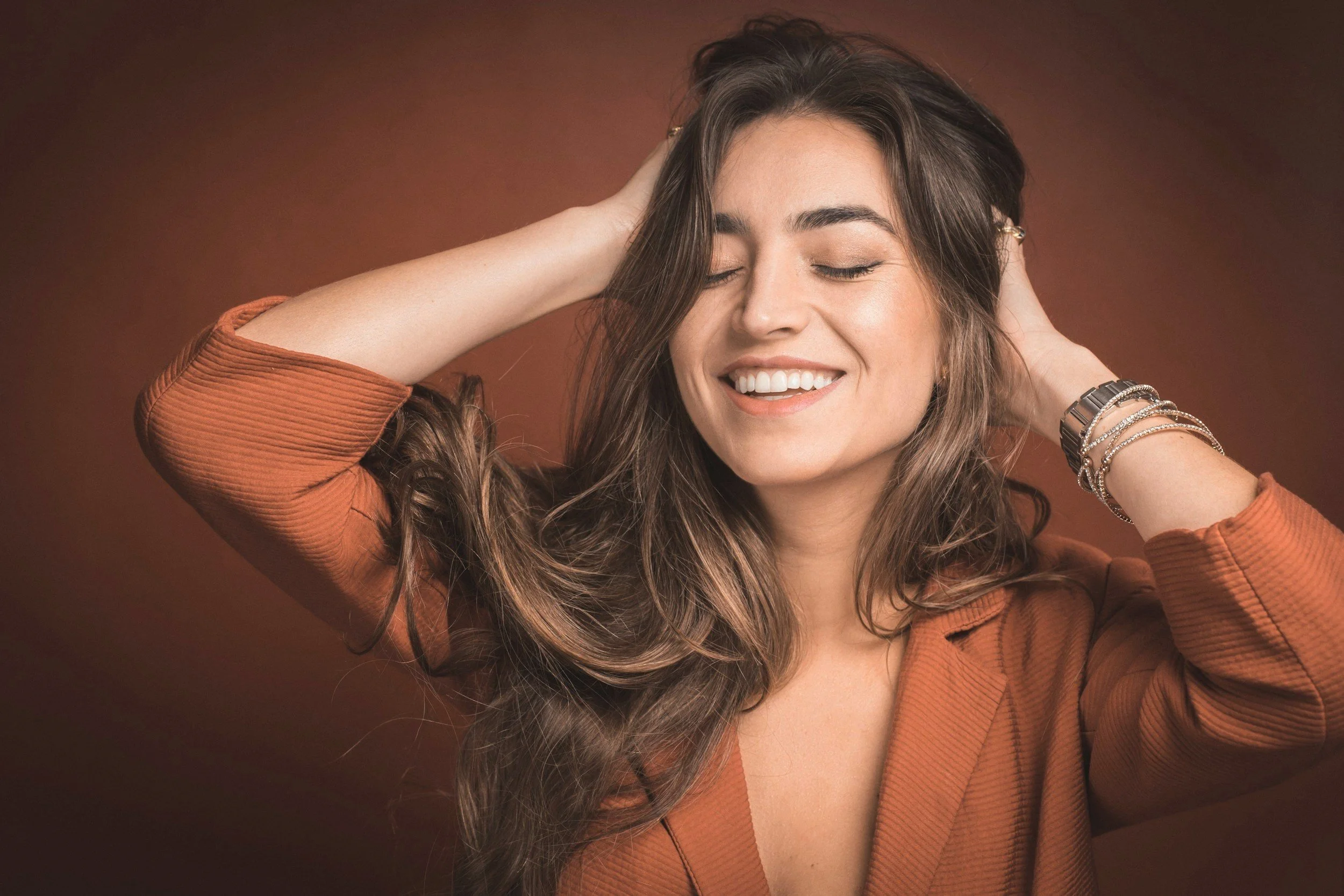 A woman with long brown wavy hair smiling and closing her eyes, wearing a rust-colored top and jewelry, against a brown background.