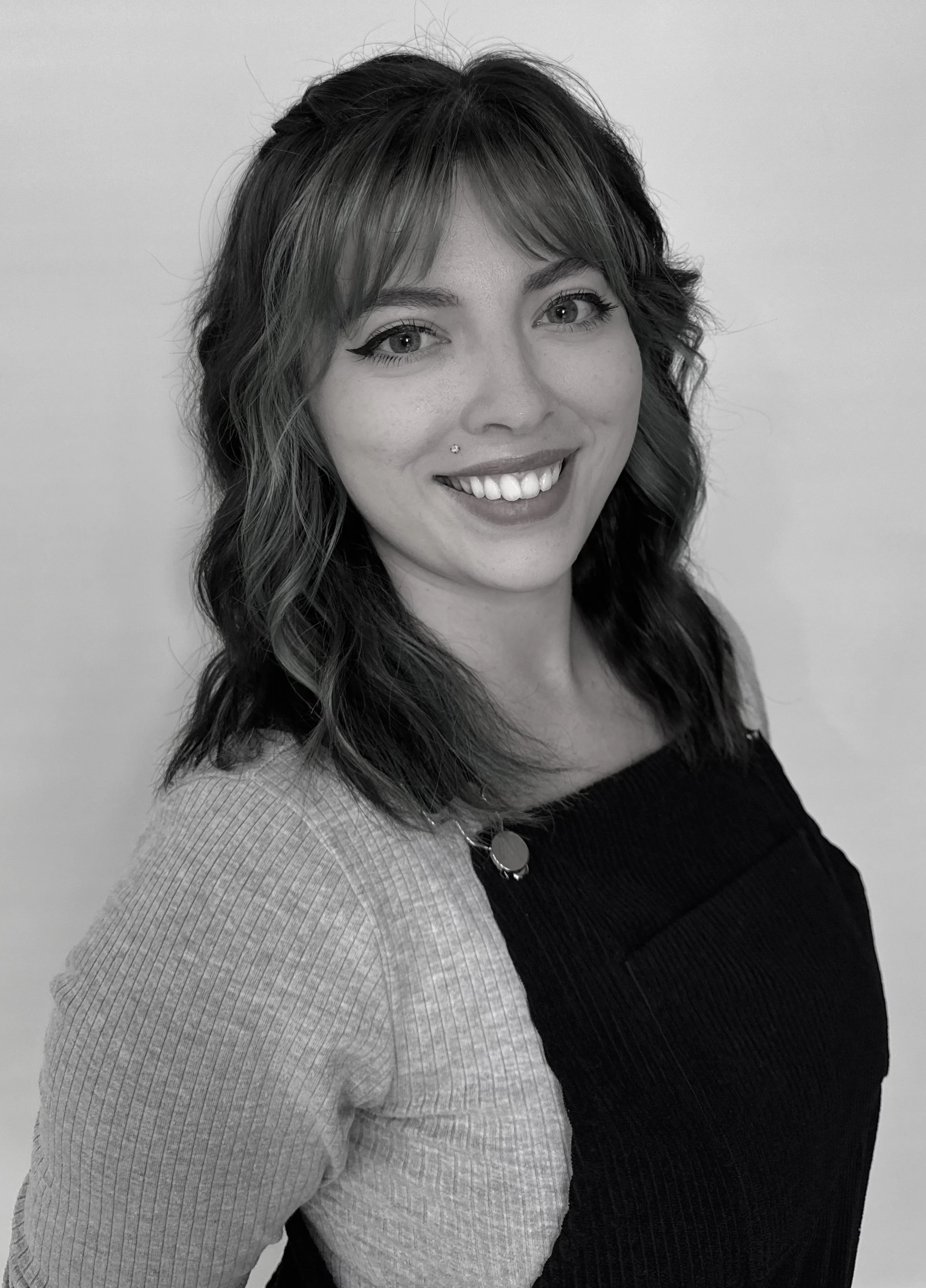 A woman with shoulder-length wavy hair, smiling, wearing a two-tone sweater with a black top and a light-colored sleeve, against a plain background.