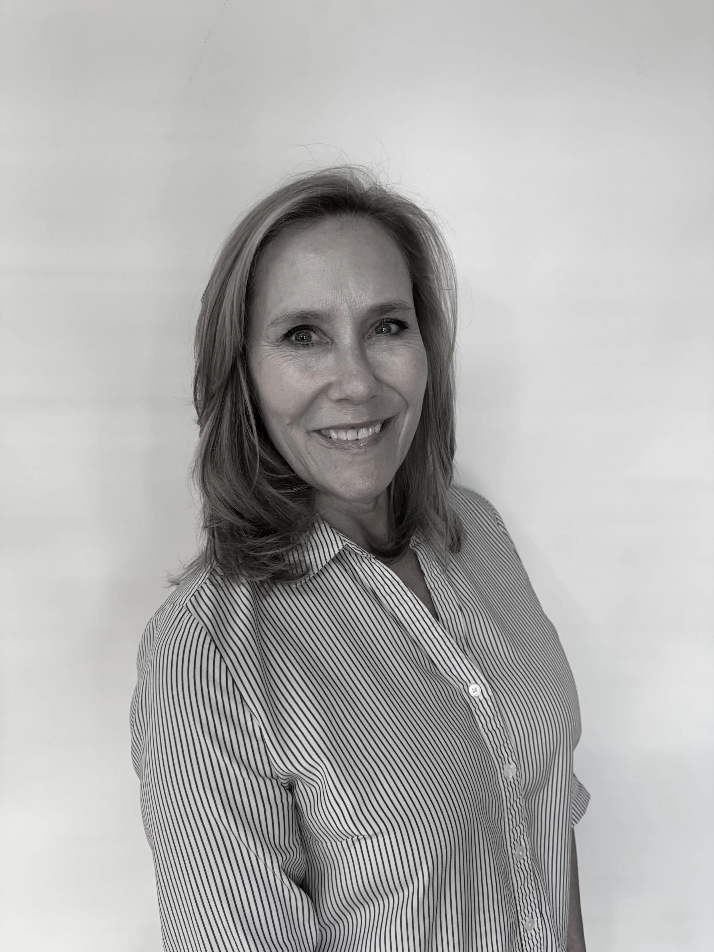 Black and white photo of a woman with shoulder-length hair smiling, wearing a striped button-up shirt, standing against a plain background.