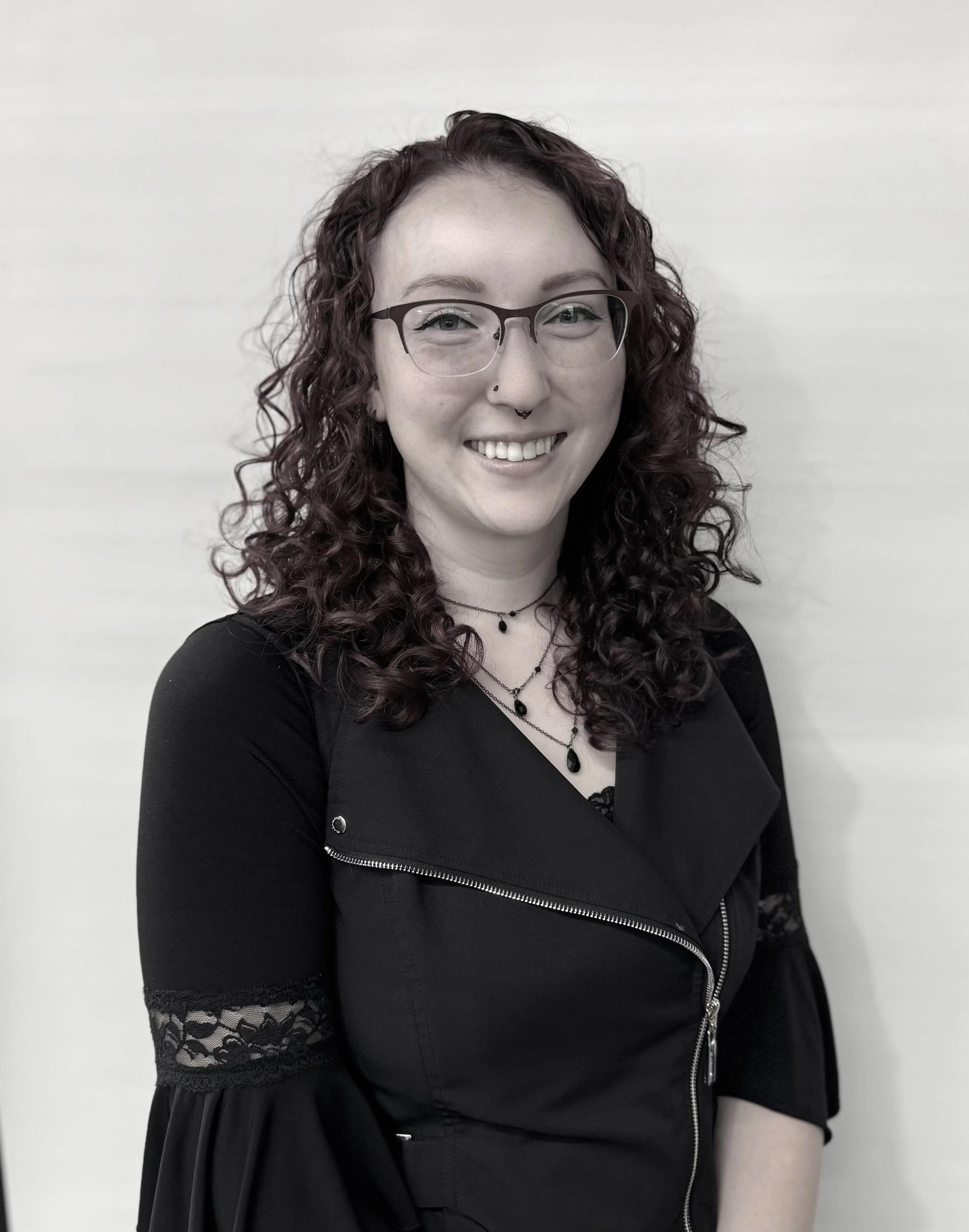 A woman with curly red hair, glasses, and a nose piercing, smiling in front of a plain light-colored wall, wearing a black outfit with lace details and layered necklaces.