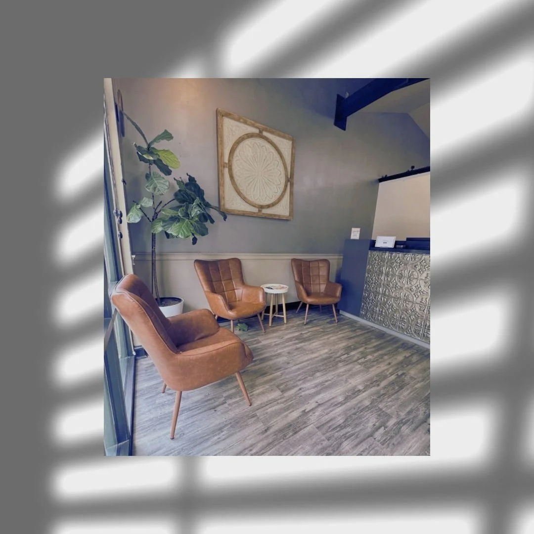 A cozy interior space with three brown leather chairs and a small round table in front of a gray wall, decorated with a large circular wall art piece. There is a tall potted plant near a glass door and the floor is covered with gray wood planks.