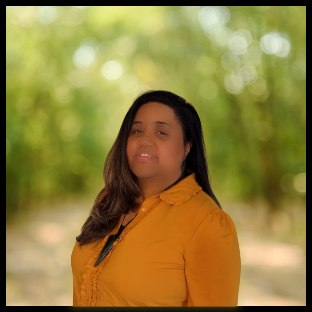 A woman with long dark hair and a mustard coloured top. She is smiling against a blurred background.
