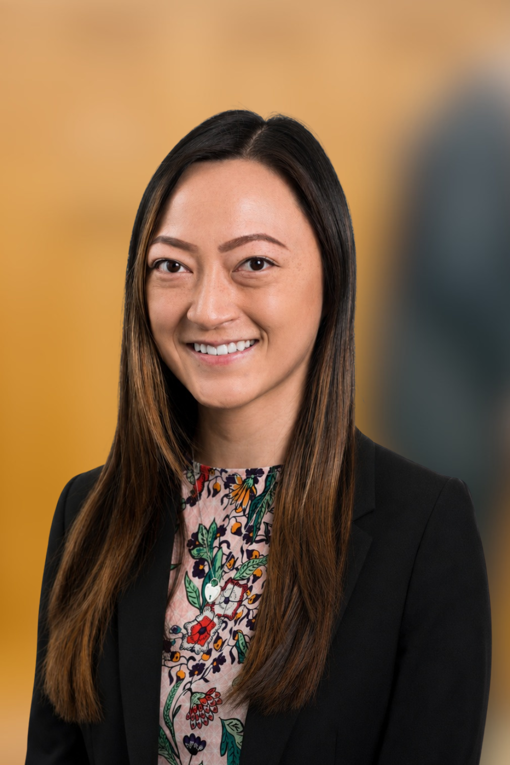 Smiling woman (Lizzie To) with long brown hair, wearing a floral pattern top with a black blazer on top,  stands in a softly blurred indoor setting.