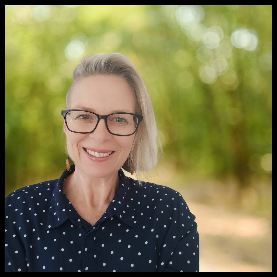 A woman with short blonde hair and glasses and smiling, wearing a dark shirt with white polka dots.