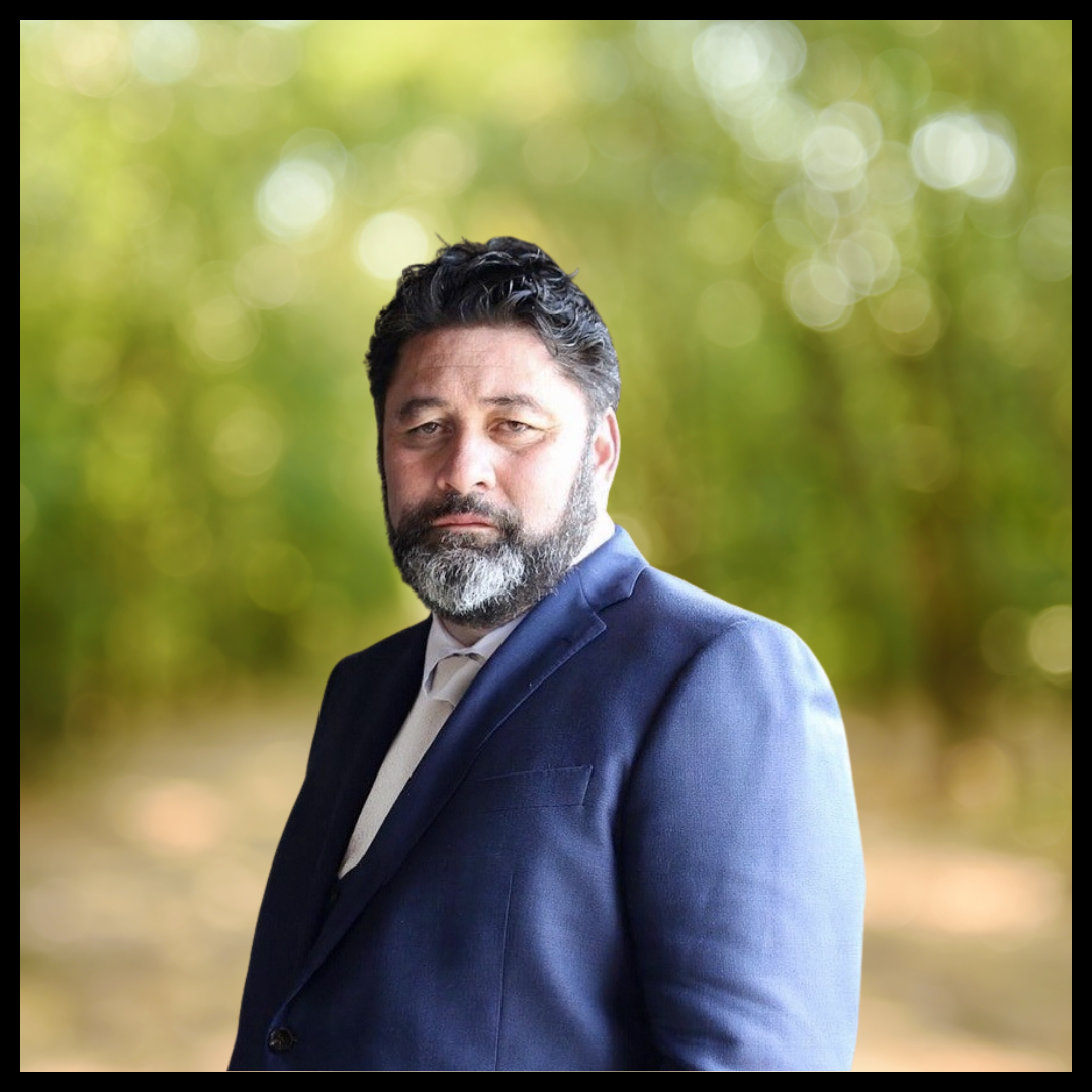 A bearded man dressed in a blue suit jacket, standing against a blurred background.