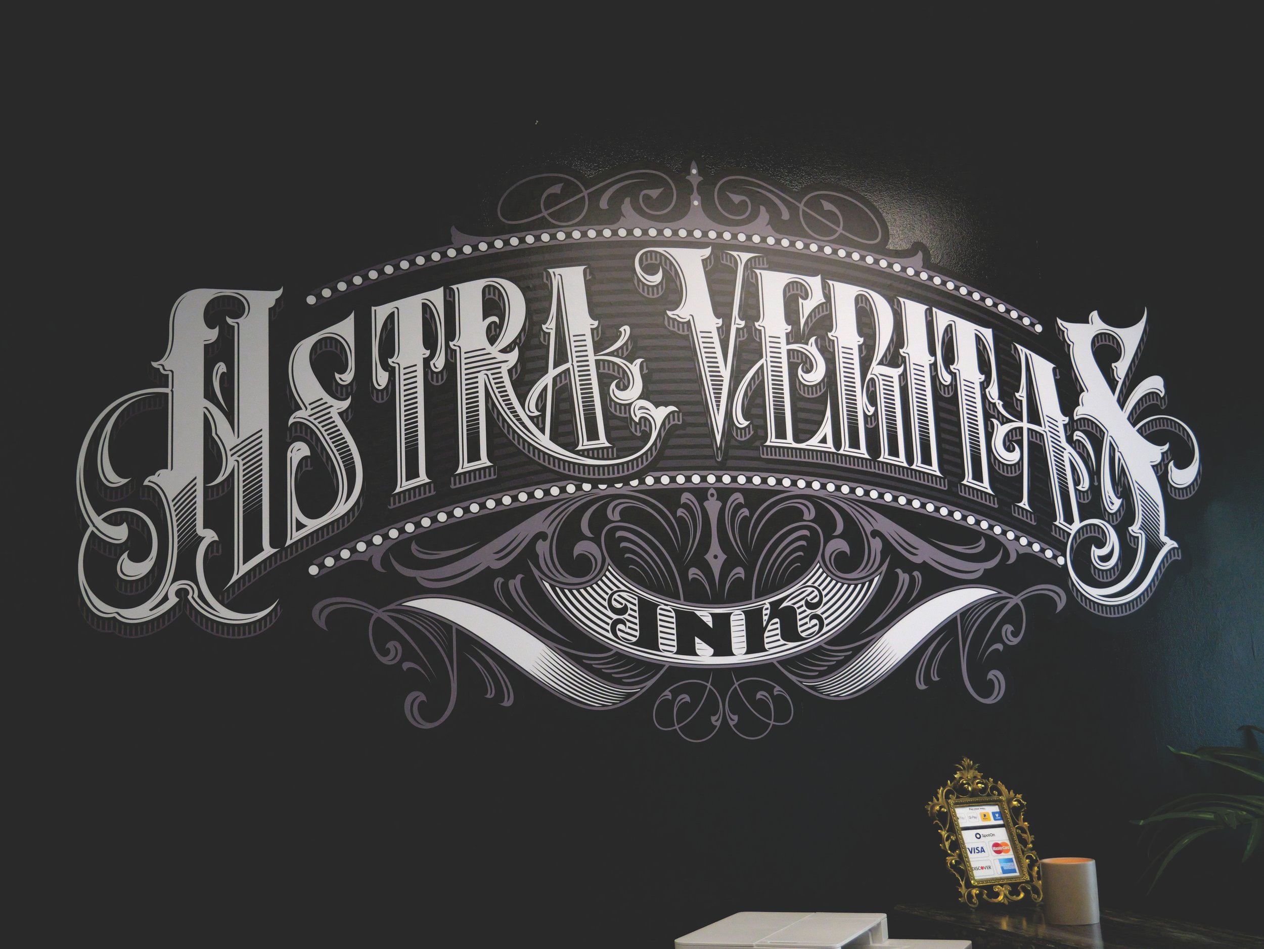 Decorative "Astra Veritas Ink" sign on a wall with ornate lettering and scrollwork.