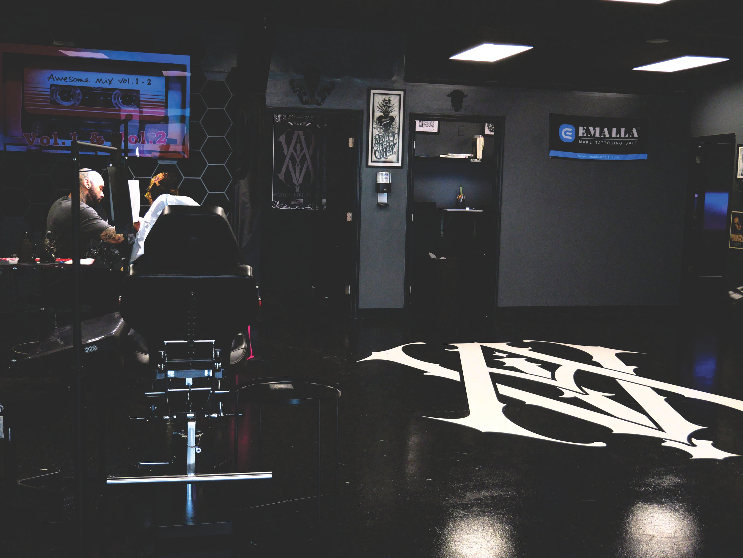 Interior of a tattoo studio with a tattoo artist working, large monogram on the floor, wall art, and equipment.