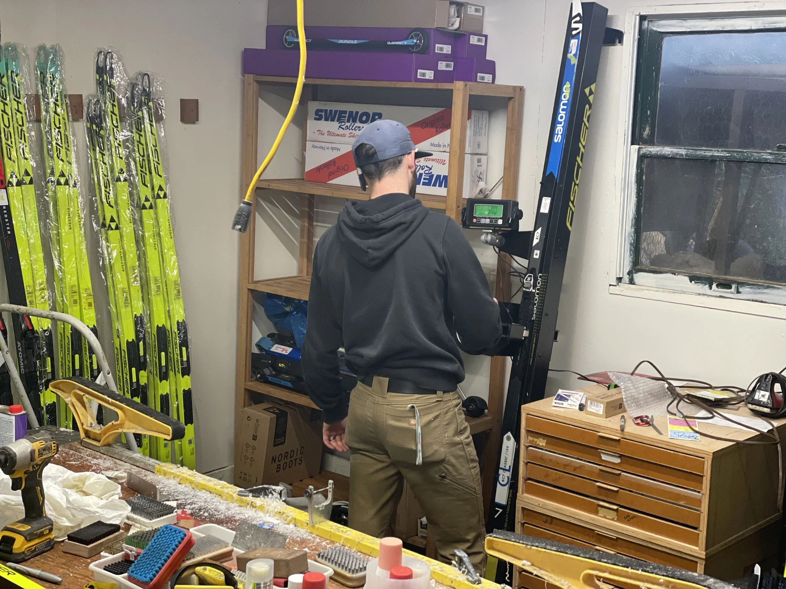 A person with a cap, black hoodie, and tan pants standing in a workshop, working on a black fitness machine. To the left, yellow and black skis are lined up against the wall. On the workbench in the foreground, there are various tools and supplies, including a drill, brushes, and small containers. A wooden cabinet with multiple drawers is on the right, and a window with a wire screen is in the background.