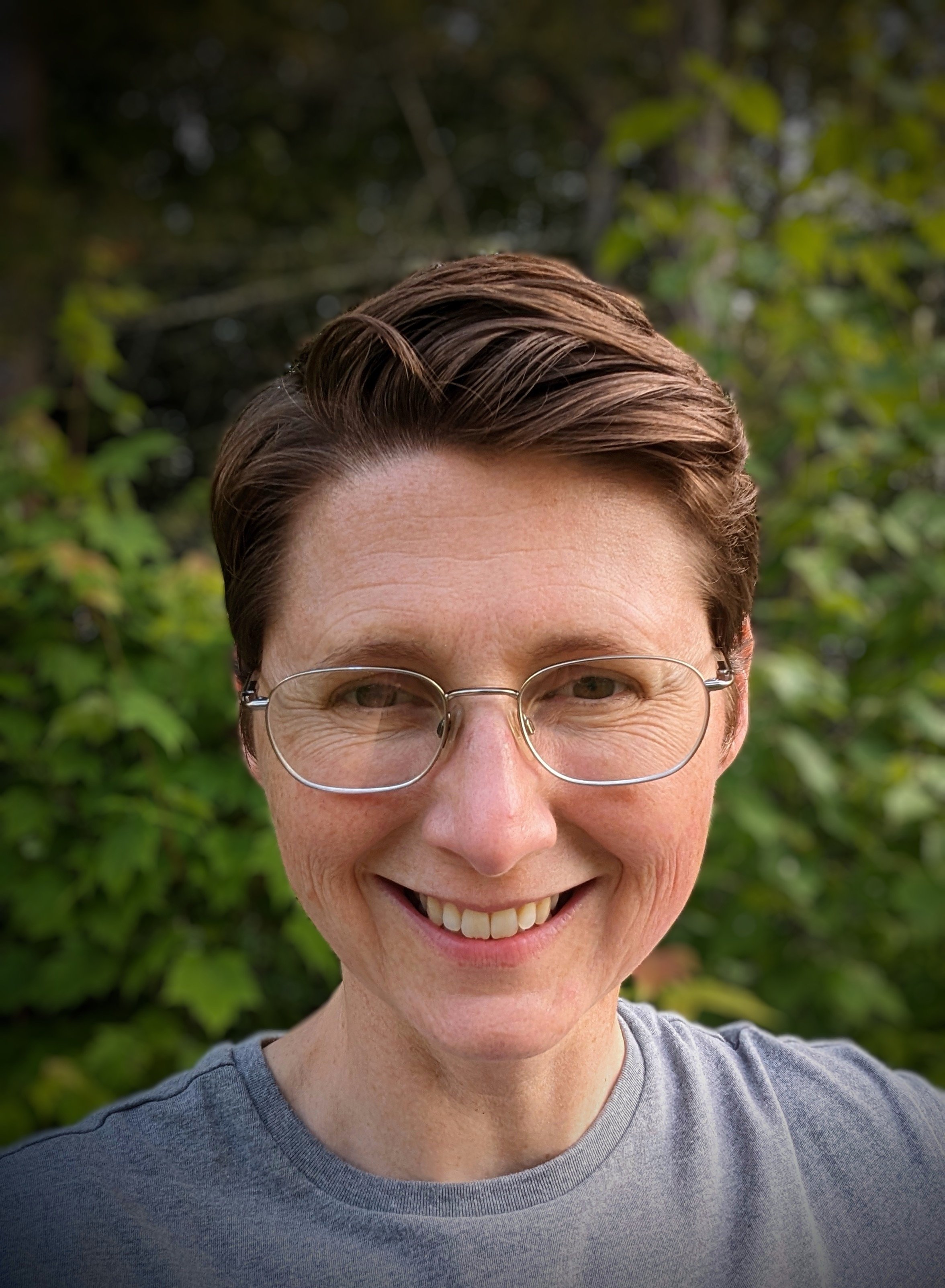 A woman with short brown hair, wearing glasses and a gray T-shirt, smiling outdoors with trees and greenery in the background.