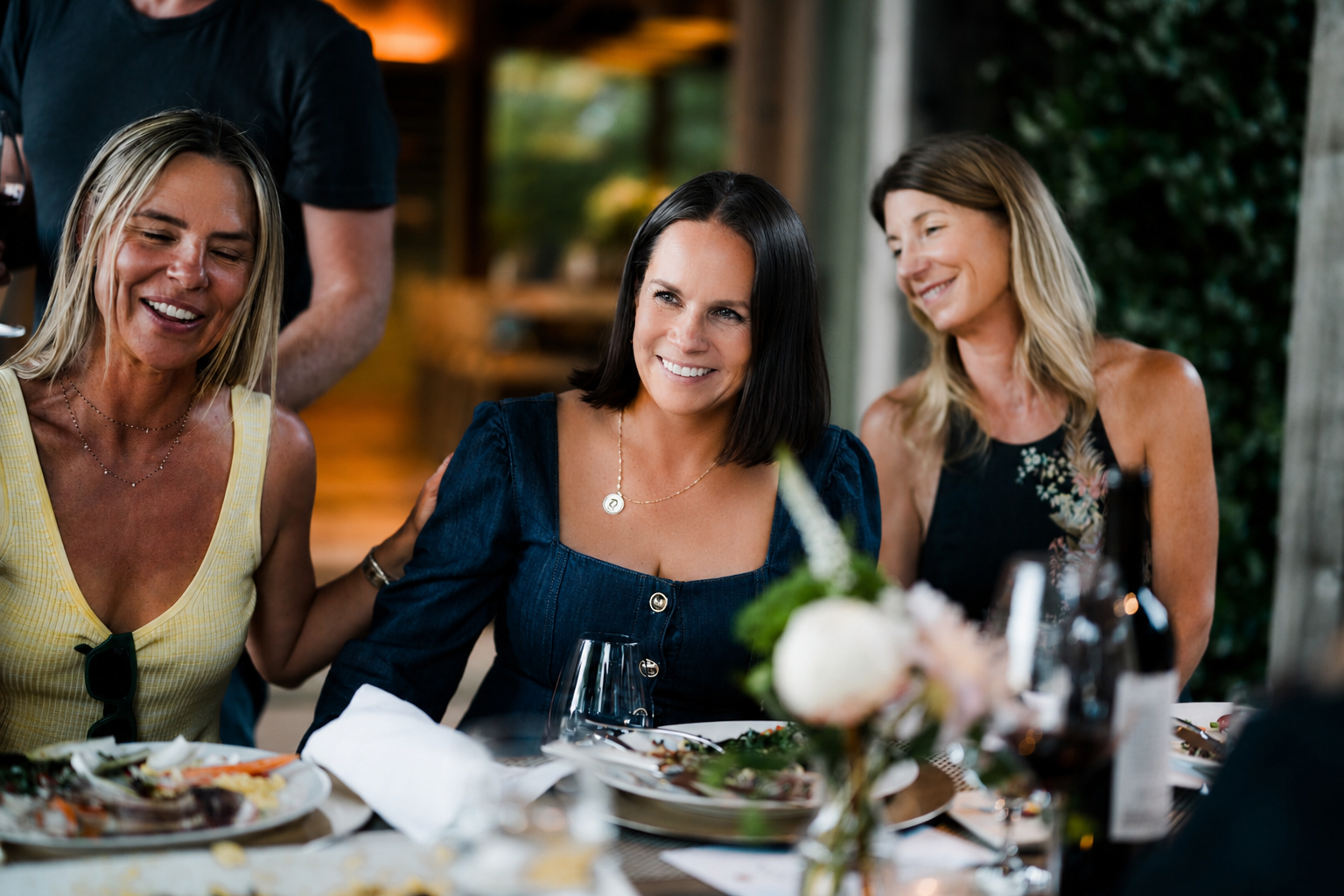 A woman with dark hair and a navy blue dress smiling at a table with food, sitting between two women. The woman to her left is blonde and wearing a yellow top, while the woman to her right has light brown hair and is wearing a black dress with floral design. They are enjoying a meal together outdoors during daytime.