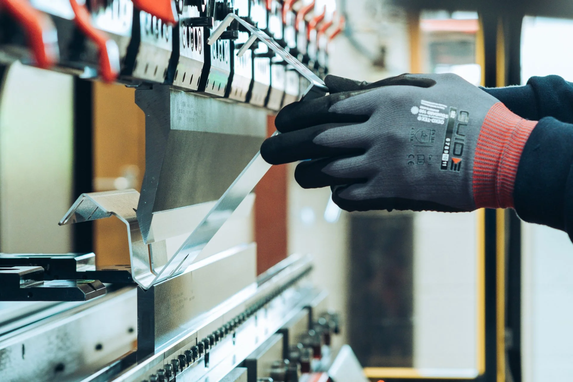A person wearing black gloves is scraping or cleaning a metal panel in a hardware store or workshop. The panel is part of a retail display with multiple compartments, possibly holding small tools or hardware items, mounted on a wall or stand. The person's hand and forearm are visible, and the background is slightly blurred.