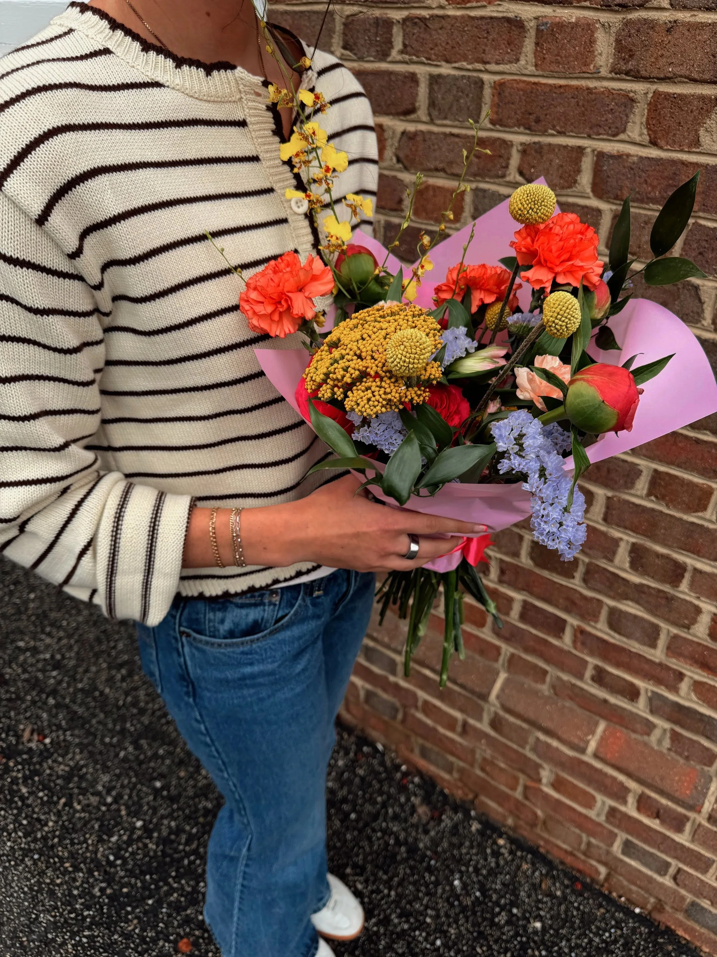 Person holding a colorful bouquet of flowers, wearing a striped sweater and blue jeans, standing against a brick wall.