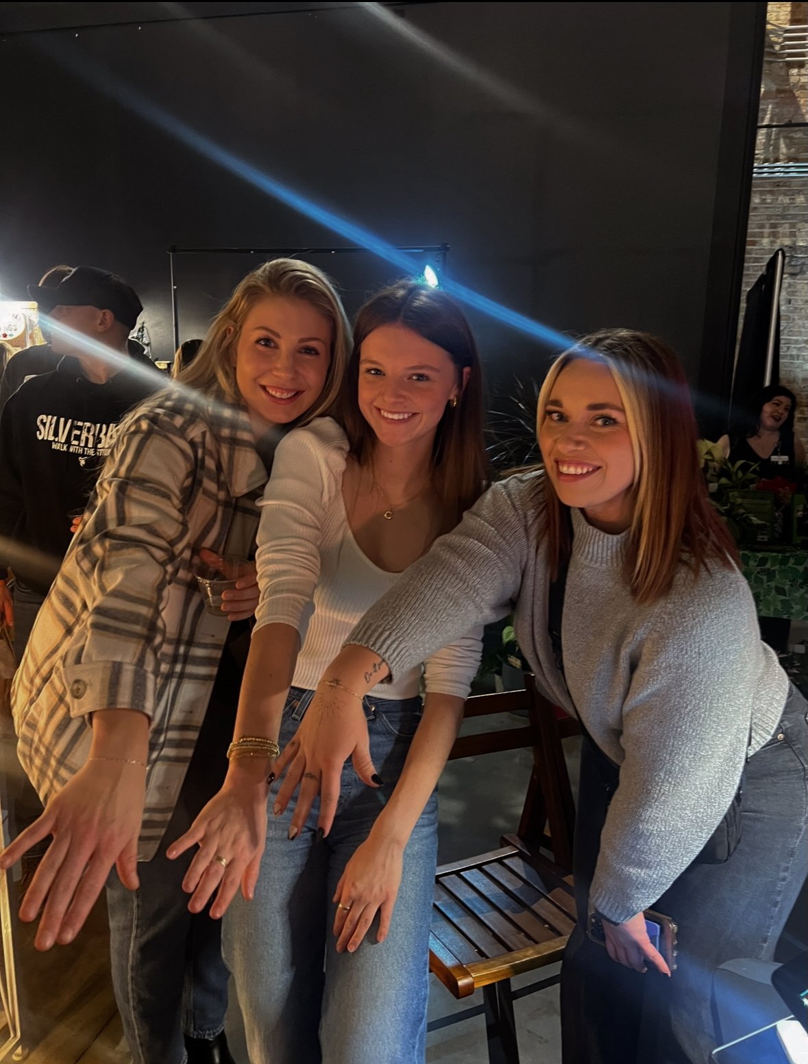 Three women smiling and reaching toward the camera at a social gathering in a dimly lit indoor space.