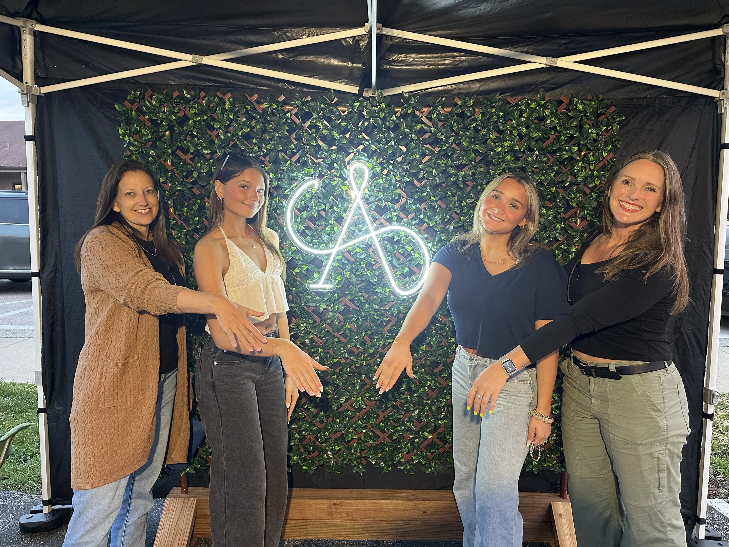 Four women standing in front of a green leafy backdrop with a neon ampersand symbol. They are smiling and have their hands extended toward the symbol. It appears to be a group photo at an event or gathering.