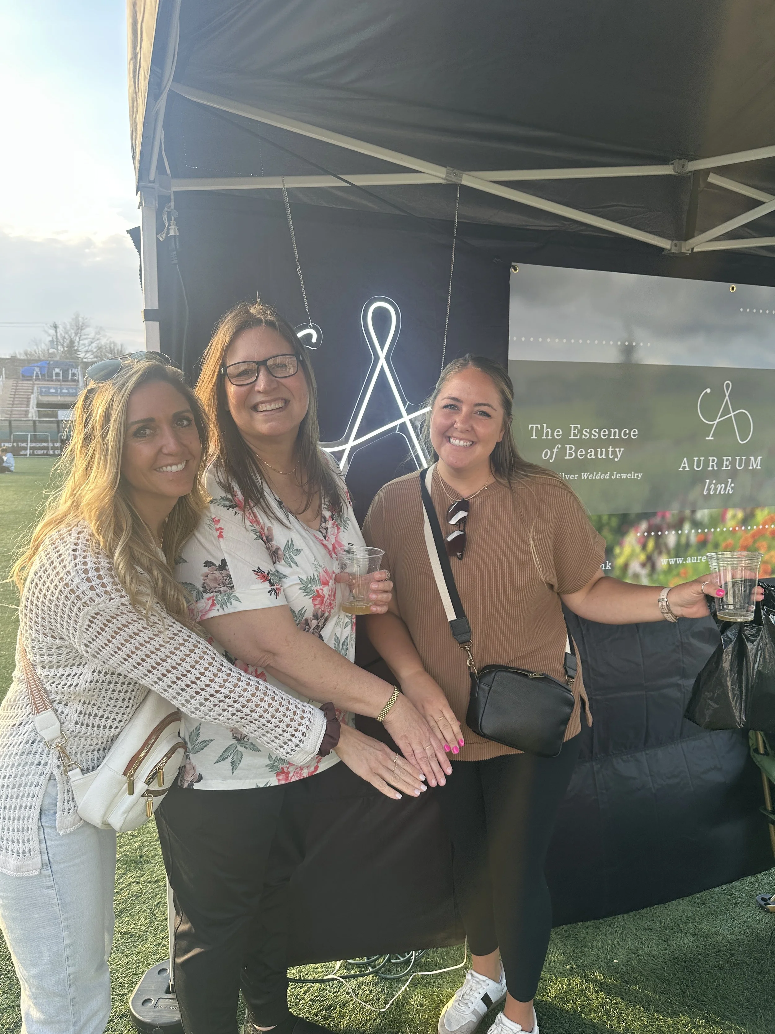 Three women standing together smiling at an outdoor event in front of a booth with a banner that reads 'The Essence of Beauty' and 'AUREUM link,' holding drinks.