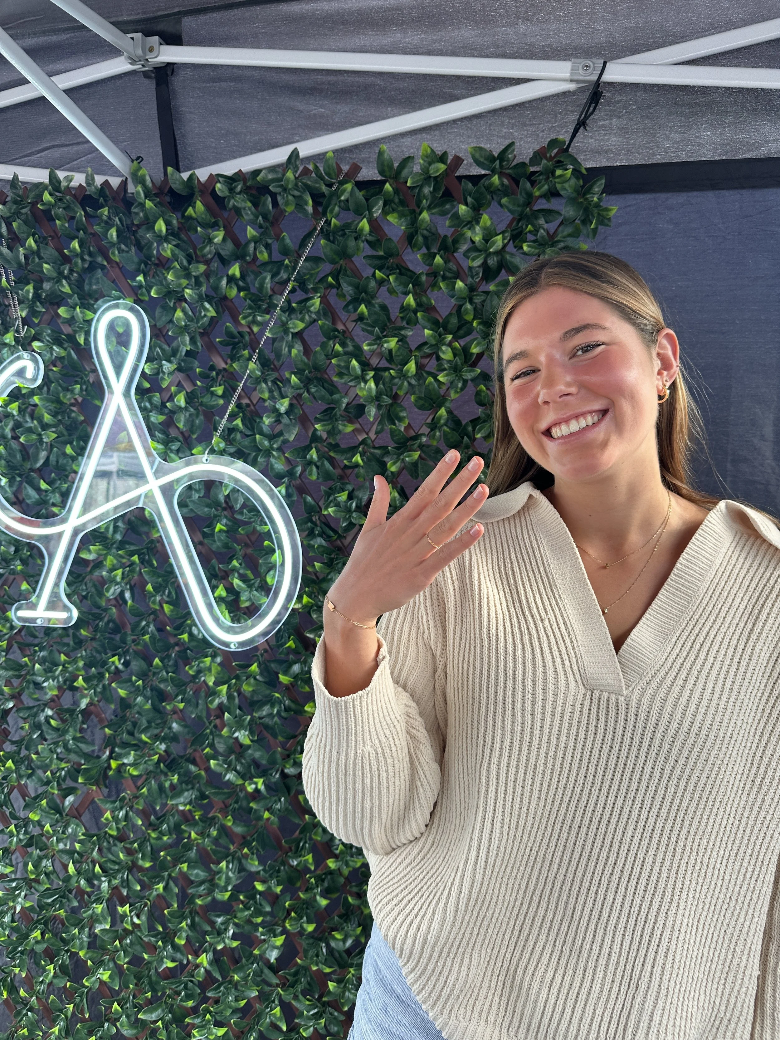 A young woman smiling, wearing a cream sweater and jewelry, standing in front of a green leafy background with a neon sign.