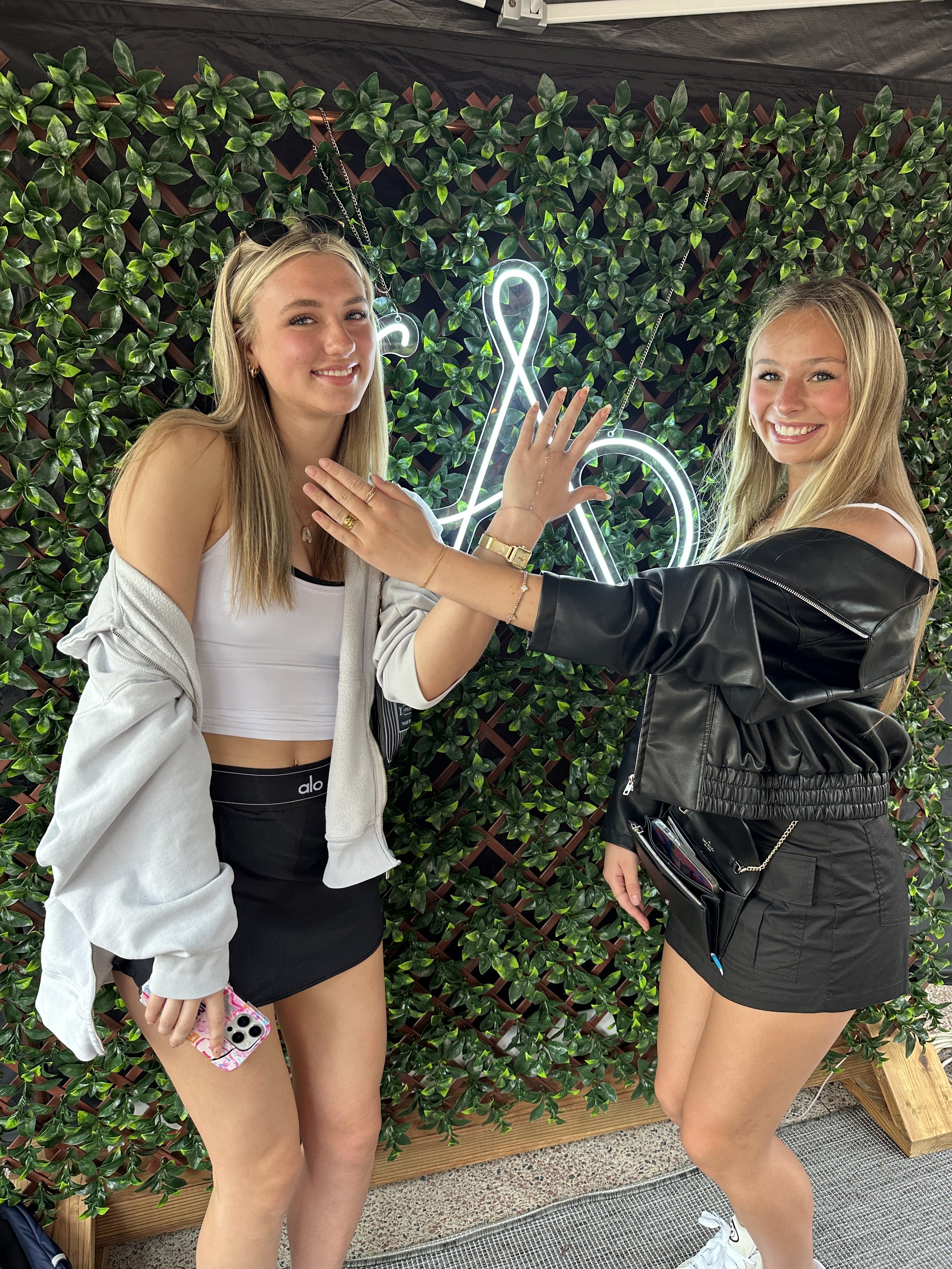 Two young women pose in front of a green leafy wall with a neon treble clef sign, giving each other a high five.