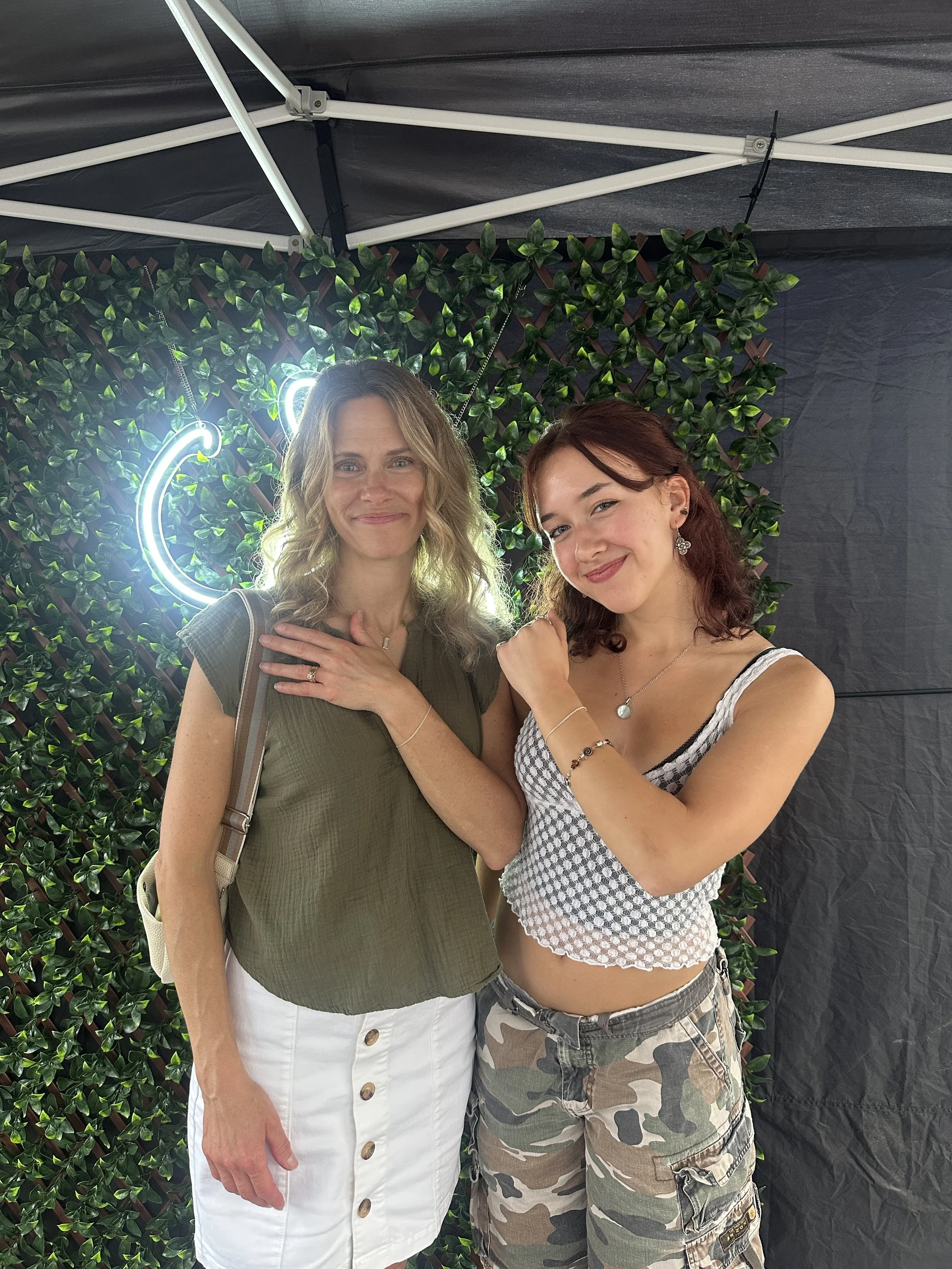 Two women standing together in front of a green leafy backdrop with neon signs, one smiling with hand over chest, the other smiling with fist raised, under a black canopy.