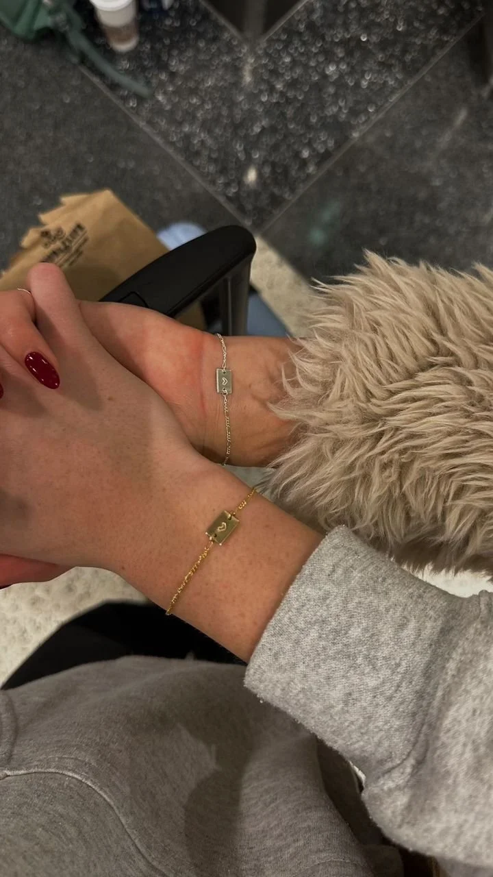 Two hands with matching gold and silver bracelets held together, one person wearing a grey sweatshirt and furry beige jacket, and the other having red painted nails. The background shows a black tiled floor and a paper bag.