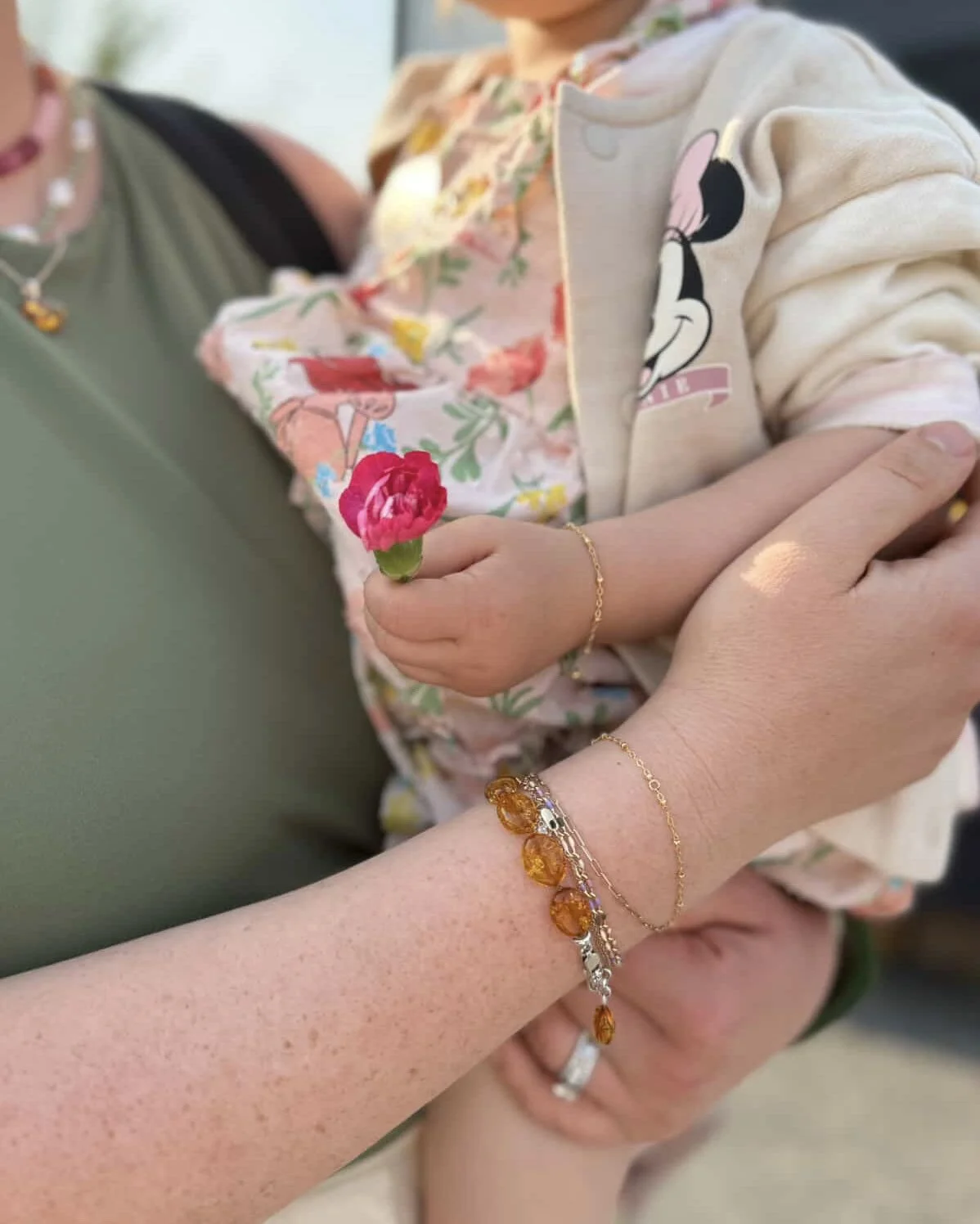 A close-up of a person's hand holding a small pink flower, with an adult and a child nearby. The child wears a Minnie Mouse jacket and the adult has jewelry, including a ring and bracelets.