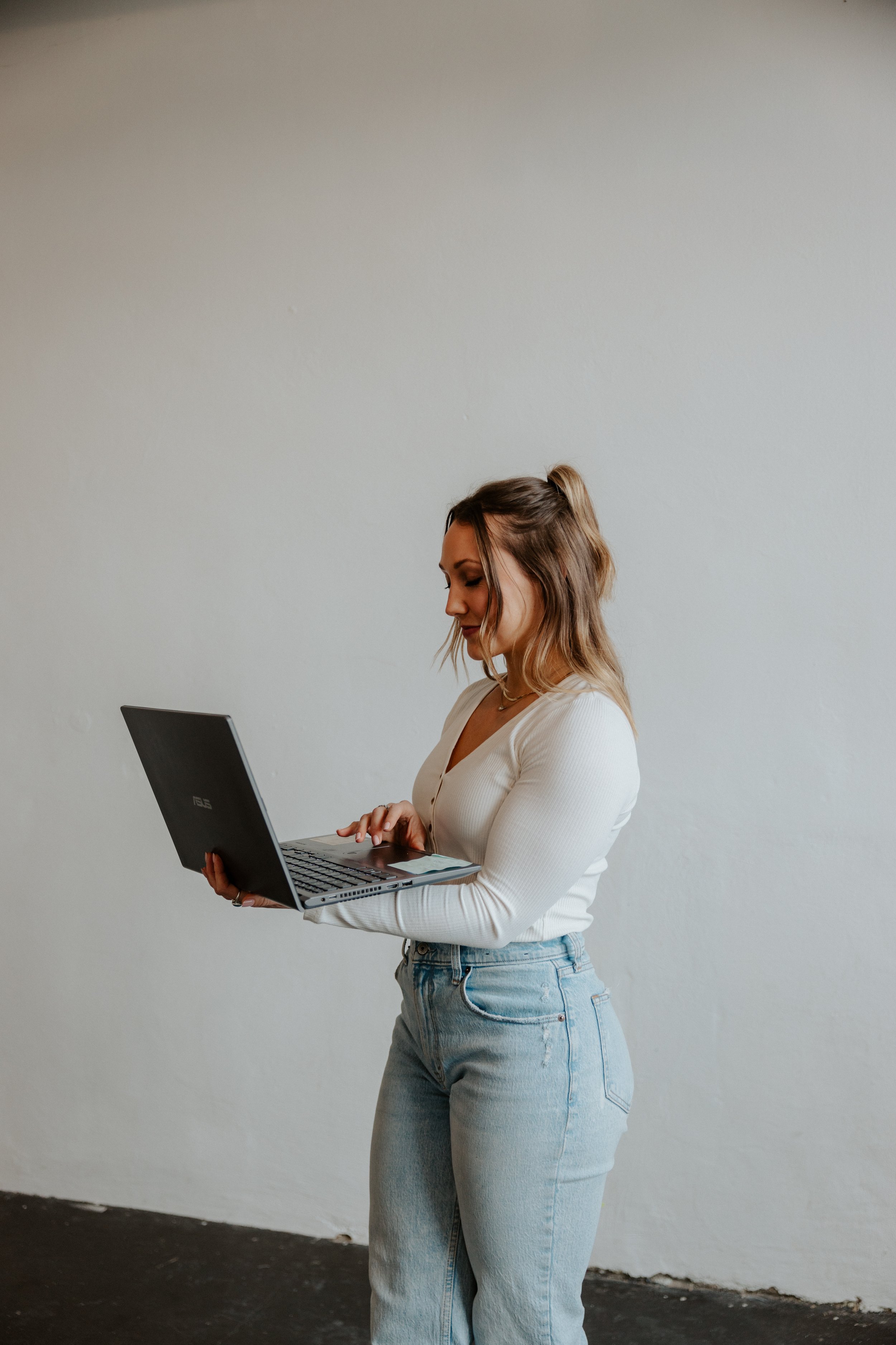 Woman with shoulder-length hair wearing a white long-sleeve shirt and light blue jeans, holding a black laptop and standing against a plain white wall.
