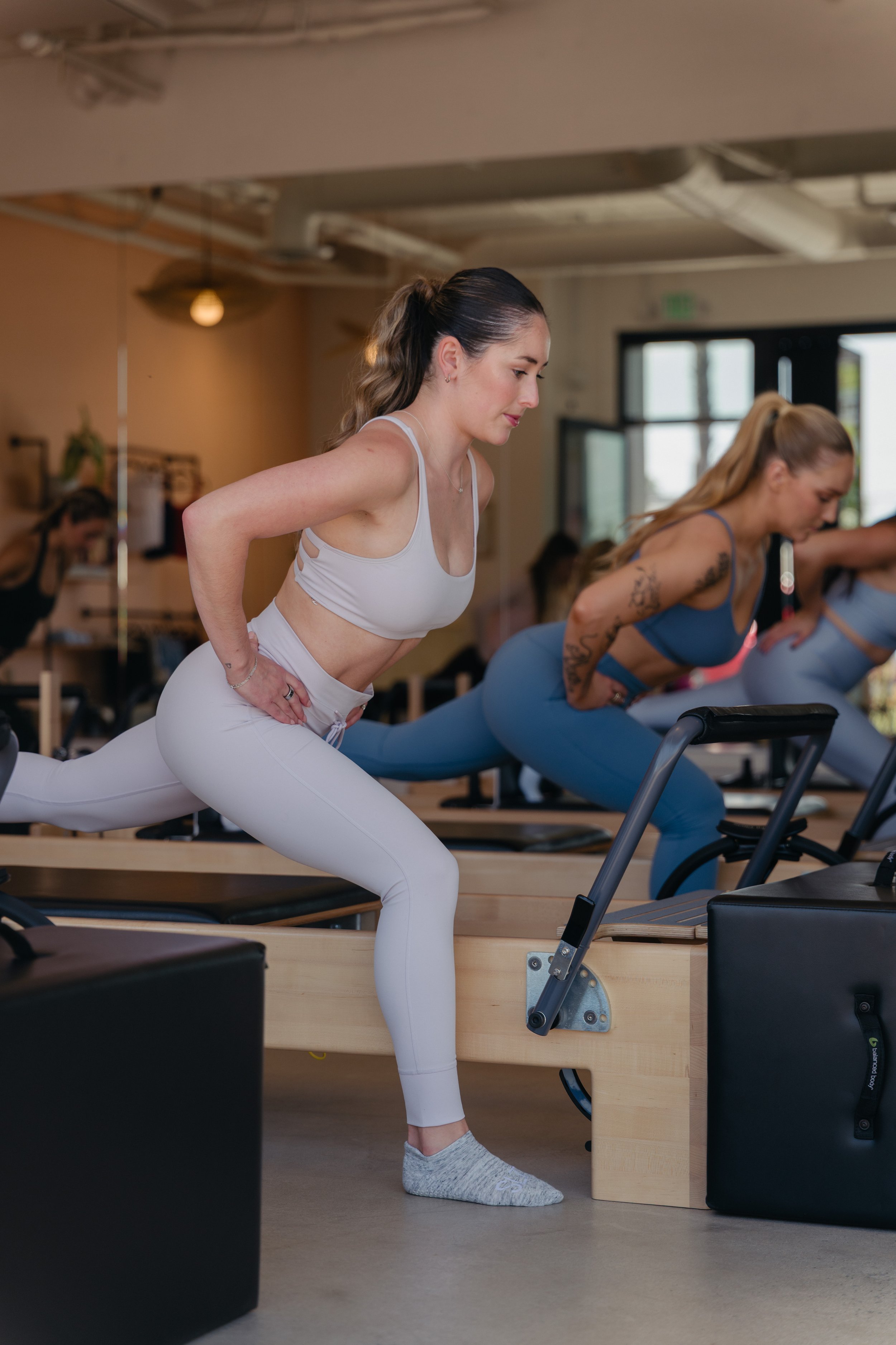 Three women practicing yoga in a fitness studio, with two women in yoga poses and one woman in a yoga pose in the foreground, all with hands in prayer position.