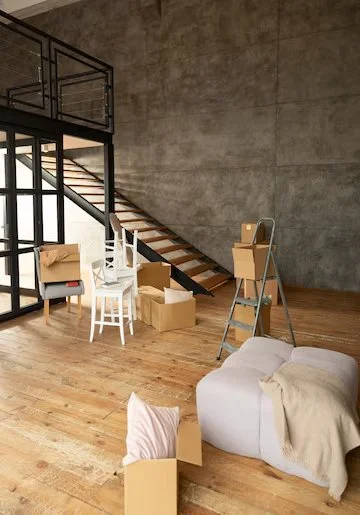 Empty living space with cardboard boxes, a white armchair, and a ladder in front of a staircase in an industrial-style loft.
