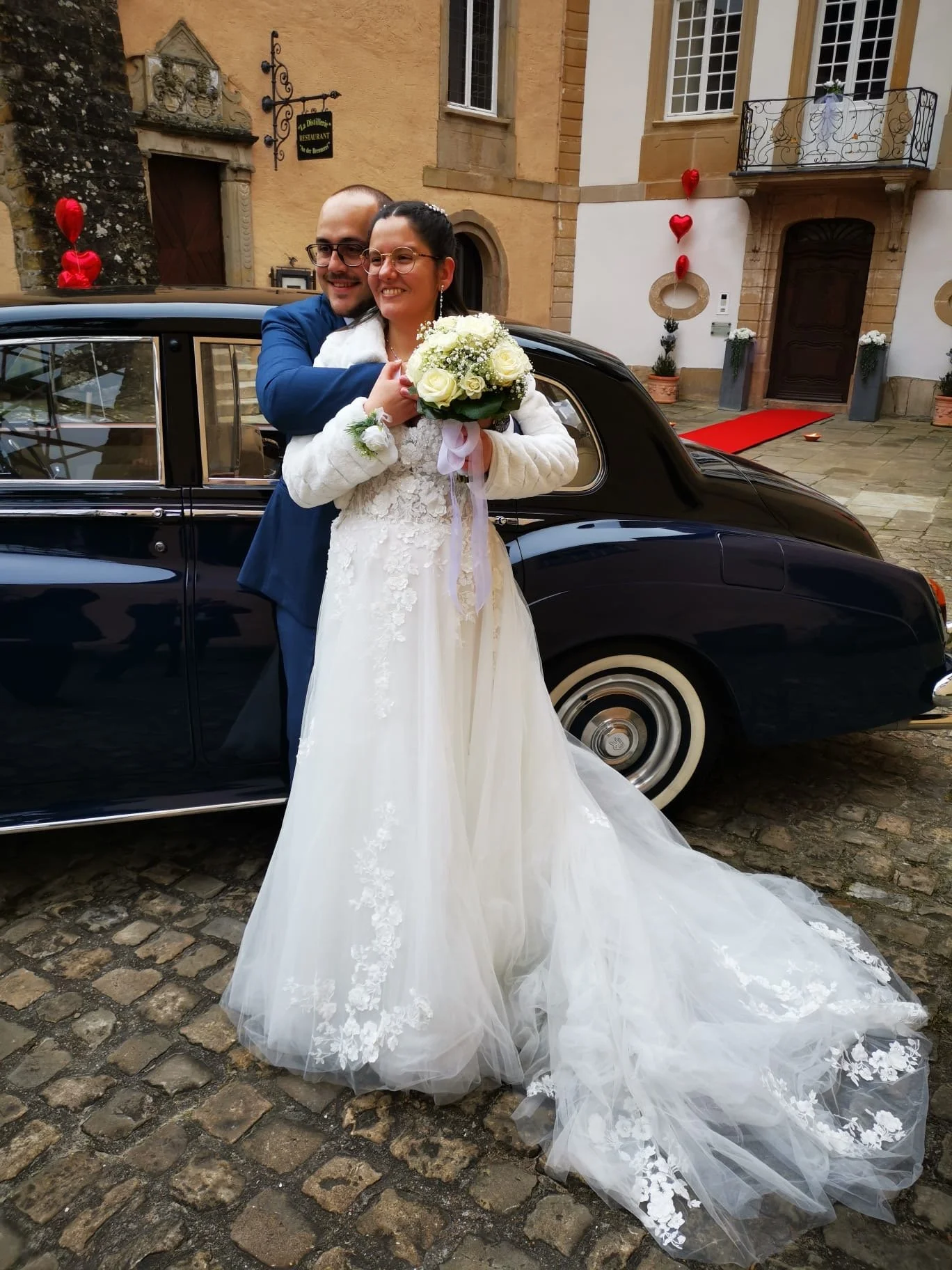 Un couple de mariés heureux posant devant une voiture noire, la femme en robe de mariée blanche et le homme en costume bleu, tenant un bouquet de fleurs.