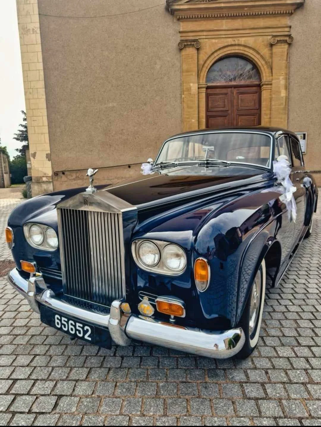Voiture de luxe noire ancienne, décorée pour un mariage, garée devant une bâtisse en pierre avec une grande porte en bois