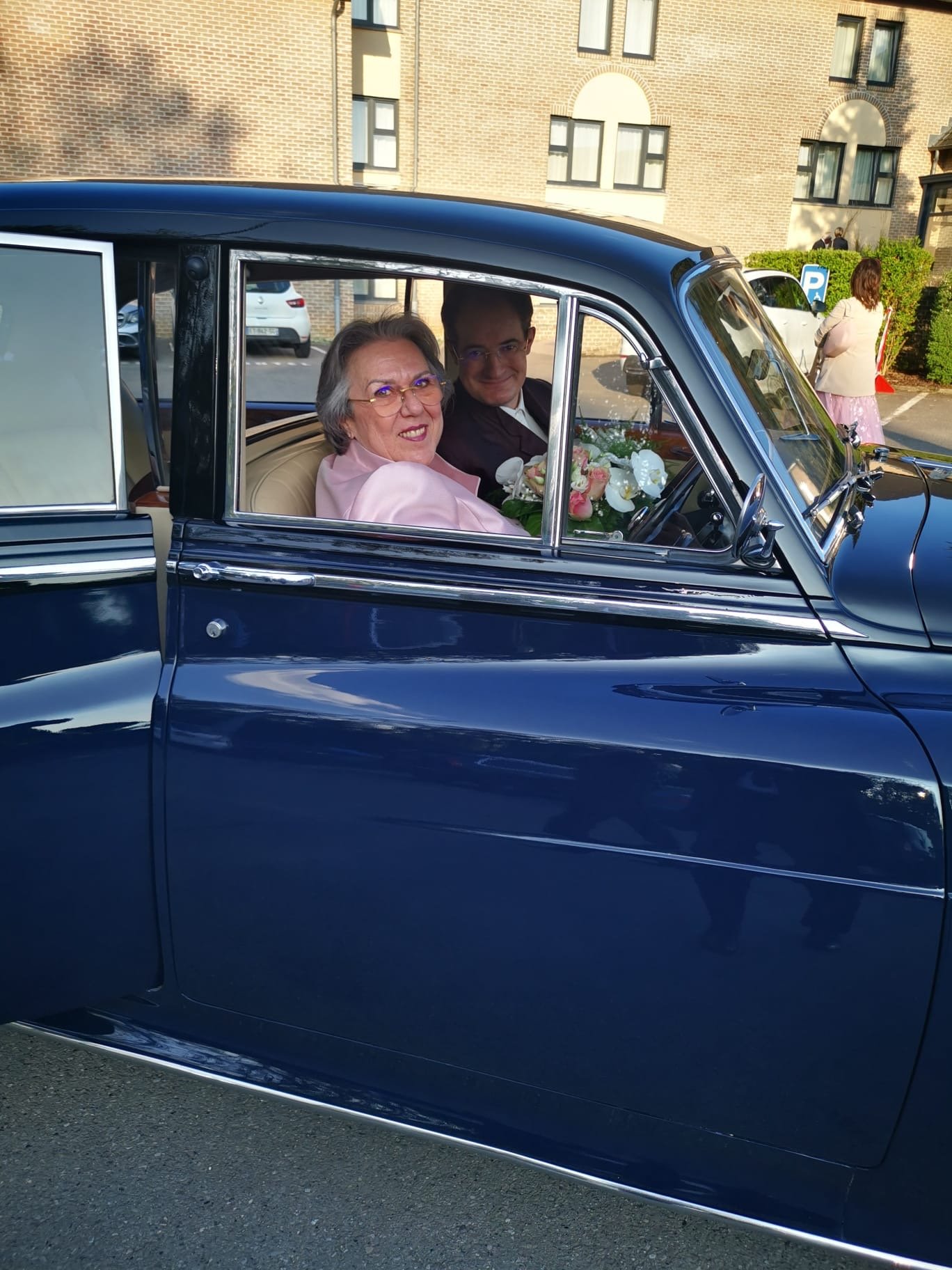 Une femme âgée et un homme à l'intérieur d'une voiture ancienne noire, avec un bouquet de fleurs, semble être lors d'un événement spécial comme un mariage, devant un bâtiment résidentiel.